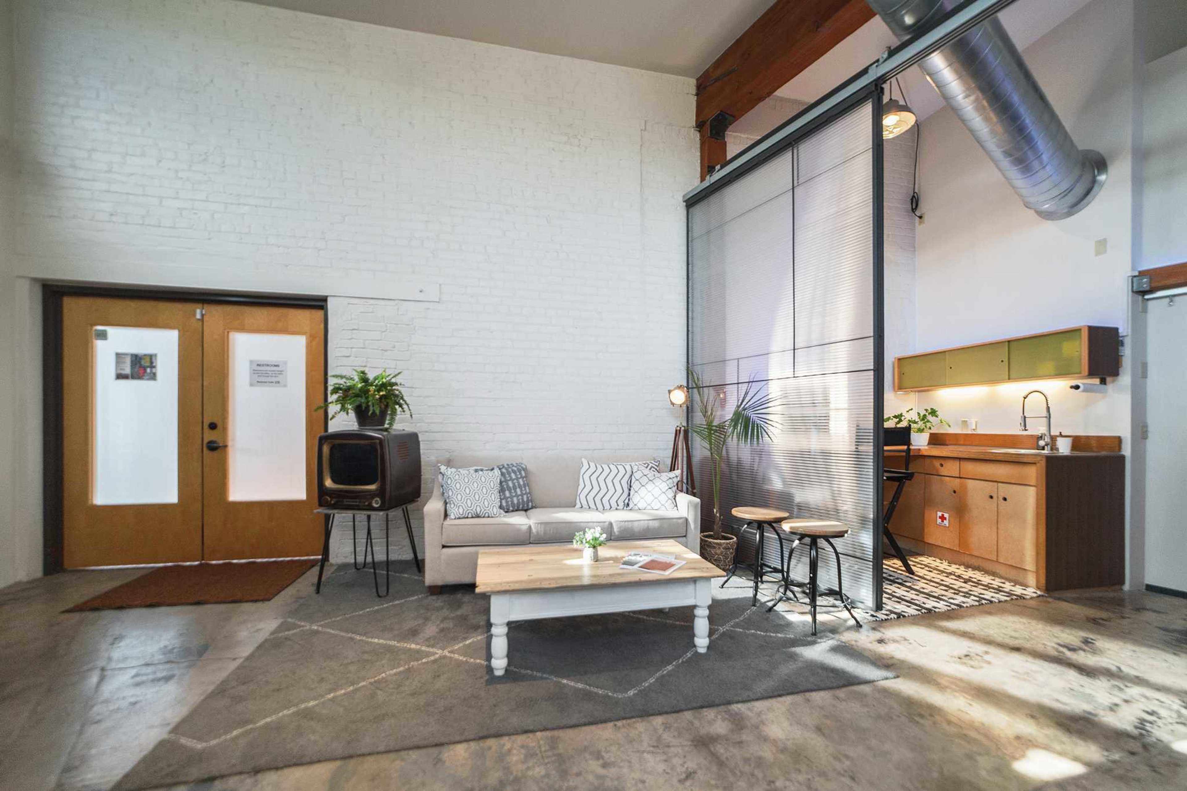 The image shows a spacious, modern living area featuring a sofa, a coffee table, and a kitchenette, with large windows and a high ceiling.