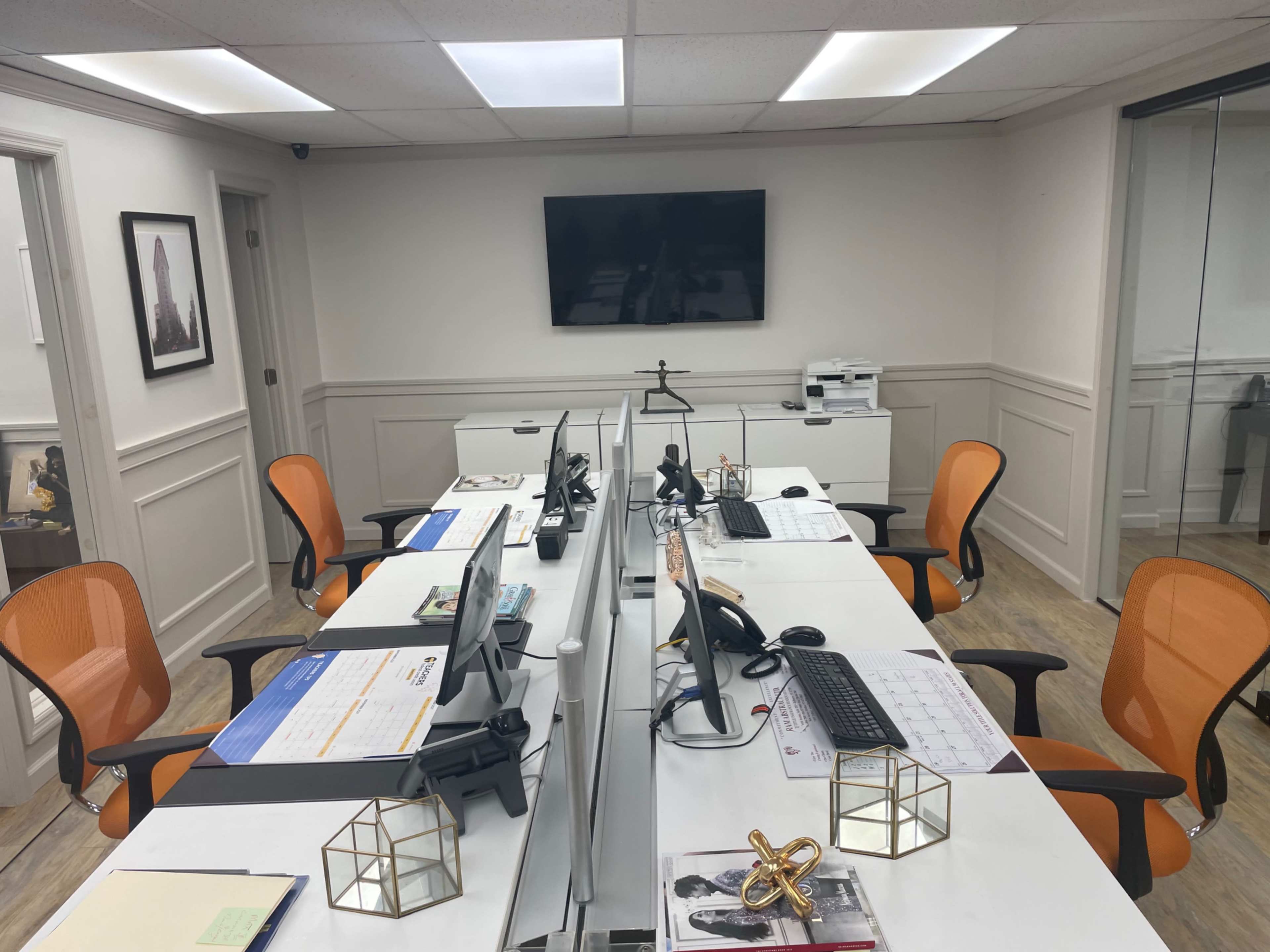 A modern office space with multiple desks, each equipped with computers, telephones, and orange office chairs, arranged in a symmetrical layout.