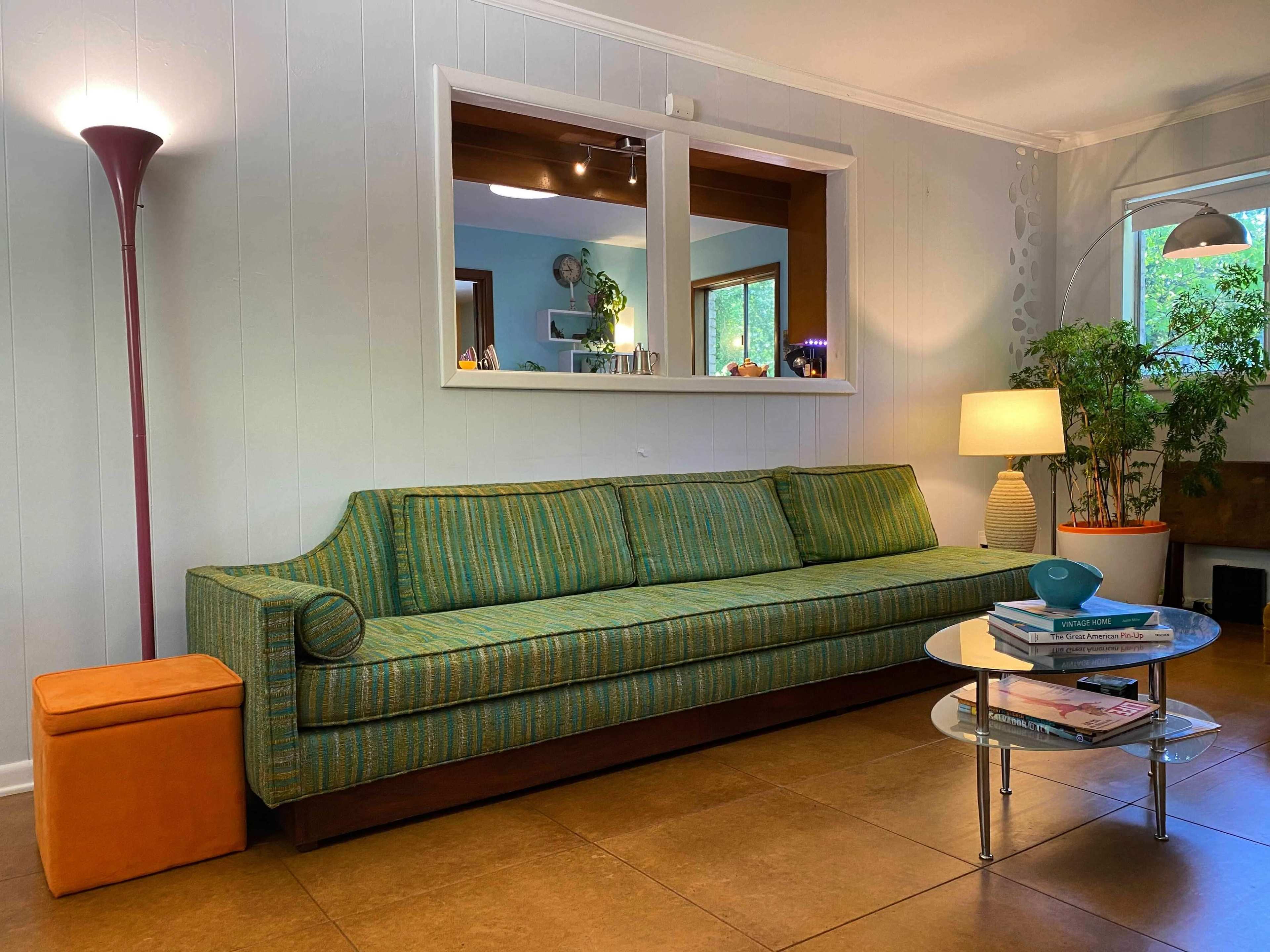 A green and blue striped sofa is positioned in a bright room with large windows, accompanied by a floor lamp, a small orange ottoman, and a glass coffee table.