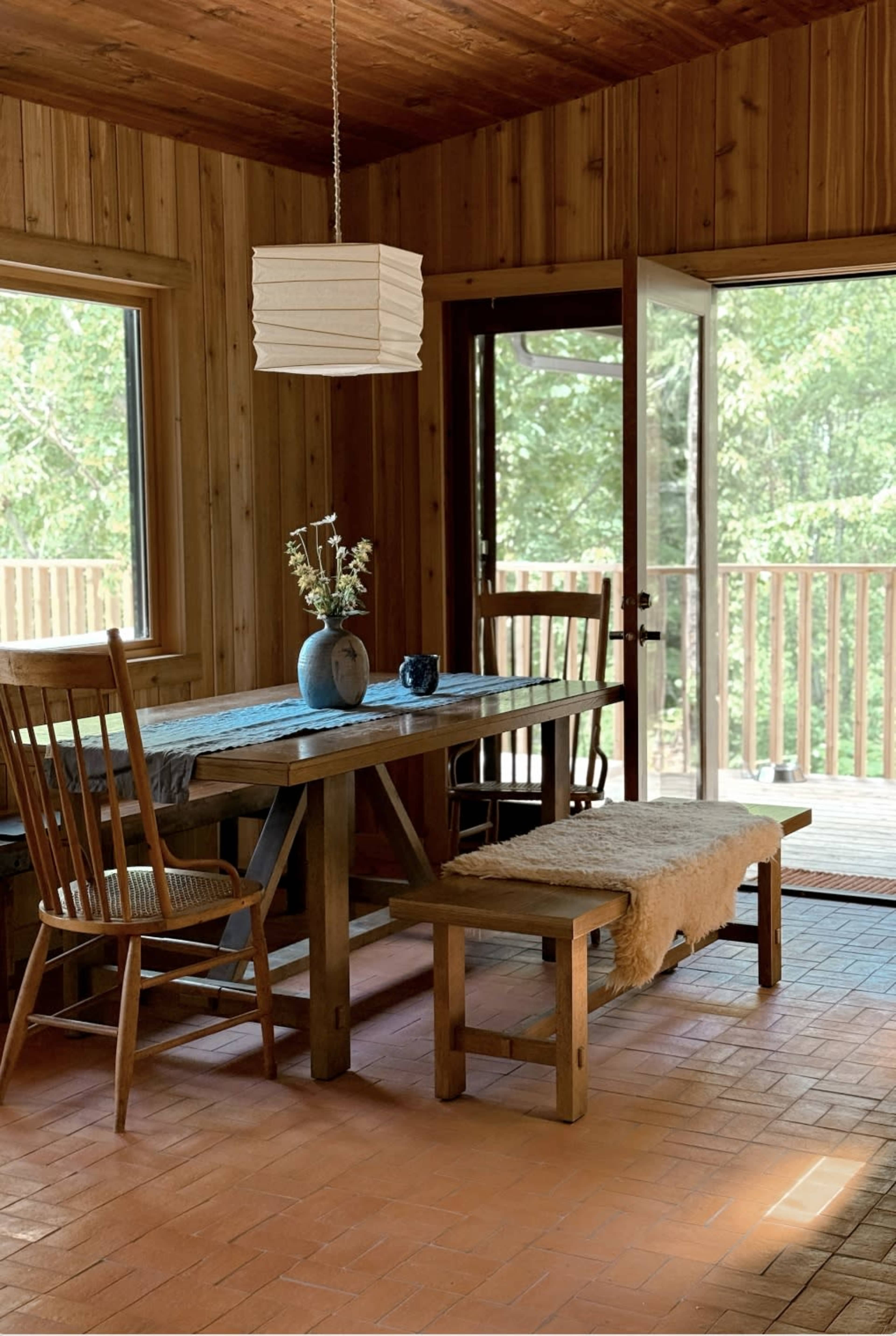 A wooden dining area features a table with a blue vase, two chairs, and a bench, with sliding glass doors opening to a deck.