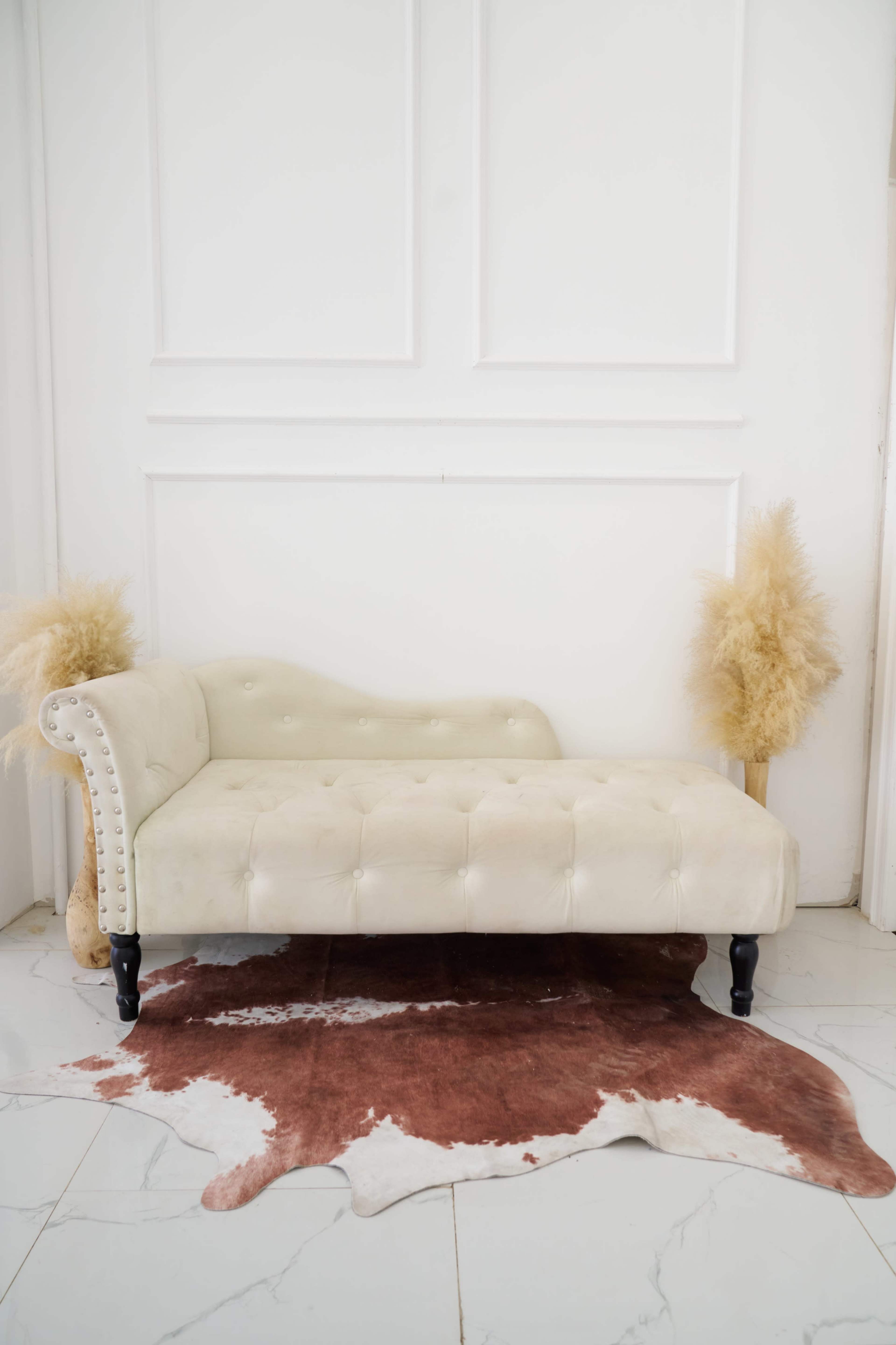 The image shows a cream-colored tufted chaise lounge placed on a brown cowhide rug, with two decorative pampas grass arrangements flanking it against a white wall.