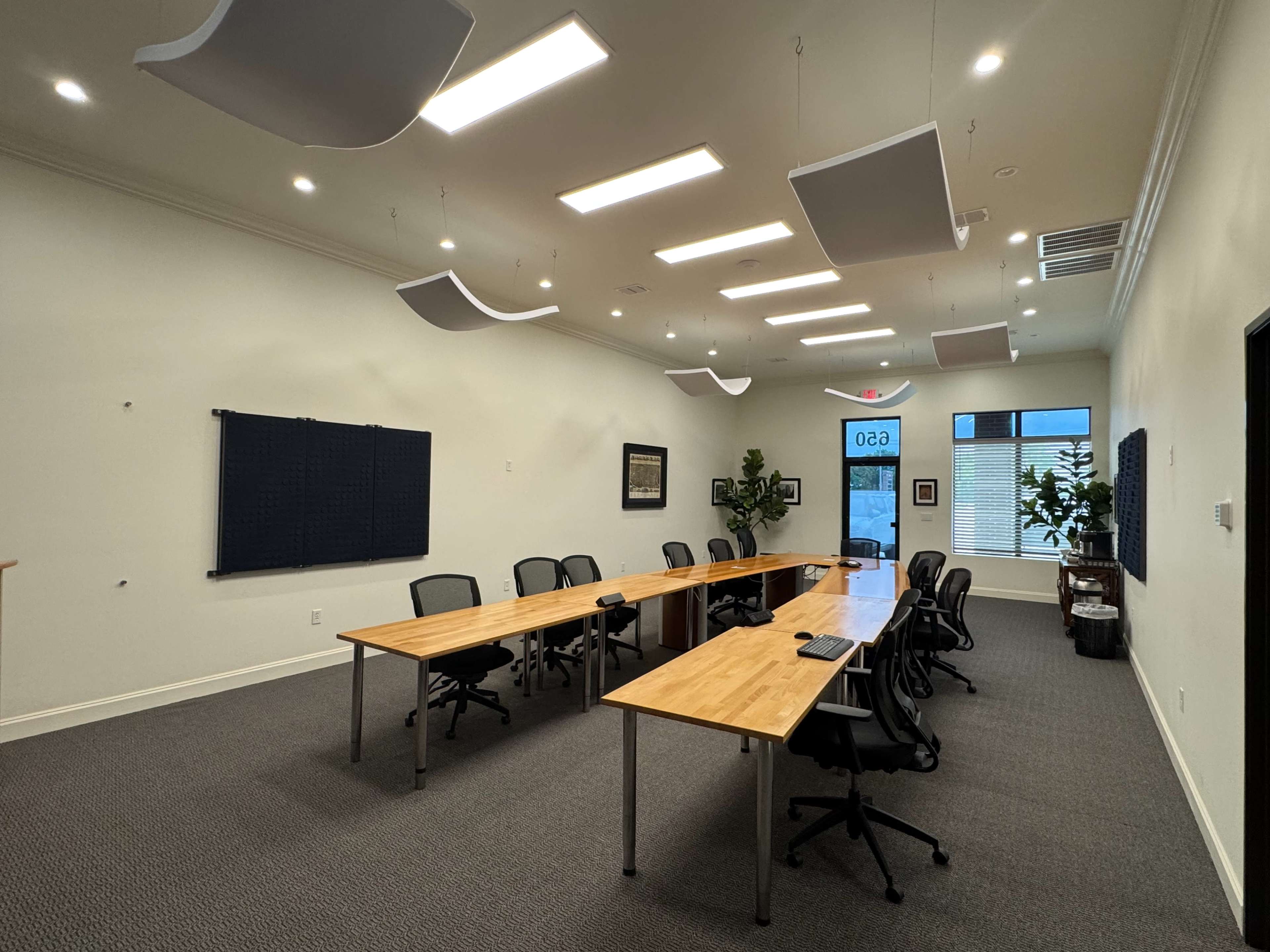 The image shows a modern conference room with a long wooden table, black chairs, and several potted plants, illuminated by overhead lights.