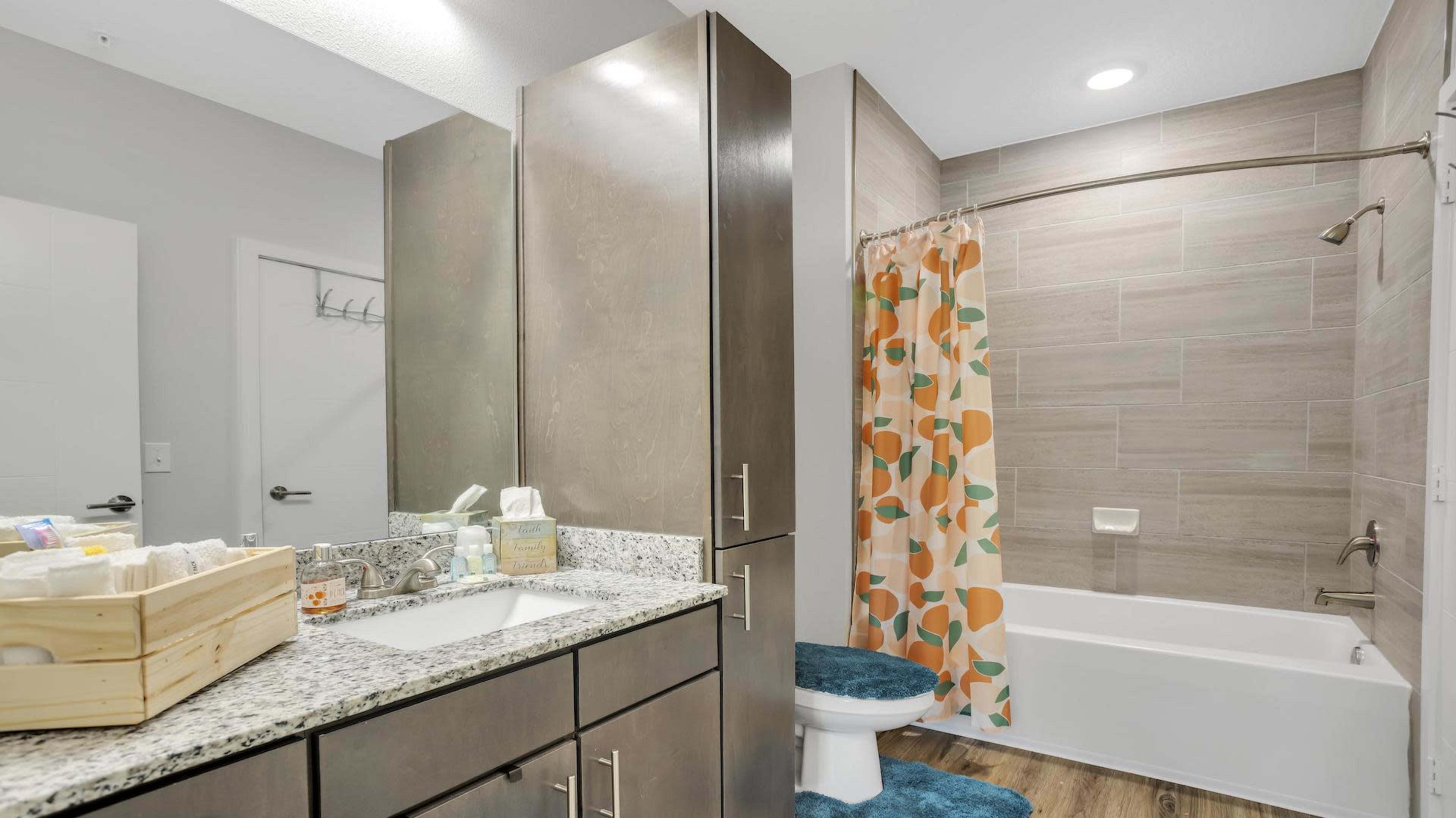 The image shows a modern bathroom featuring a bathtub with an orange and teal shower curtain, a granite countertop with a sink, and wooden flooring.