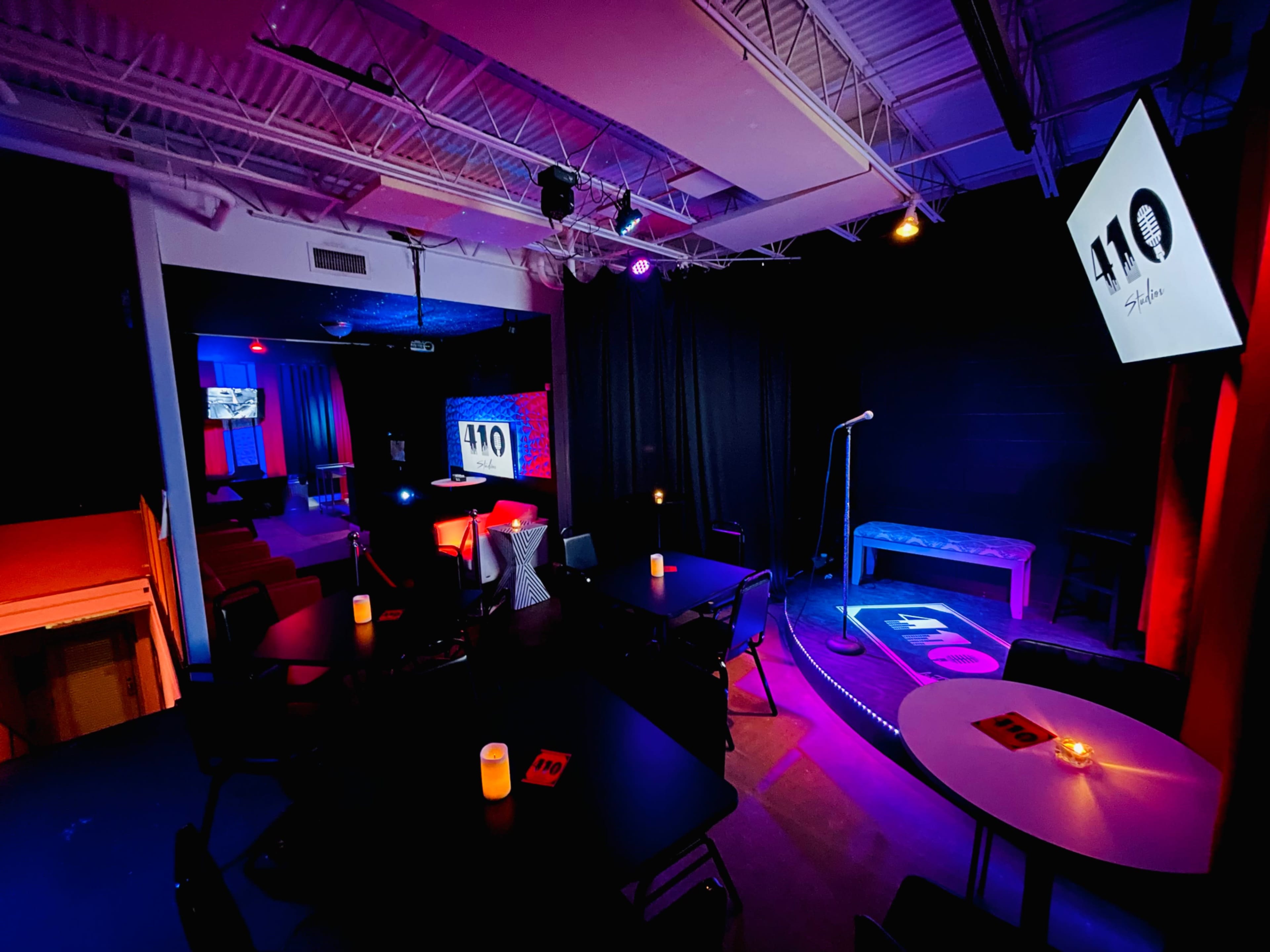 The image shows an intimate performance space with dim lighting, tables arranged for seating, a stage with a microphone, and a screen displaying the number "110."