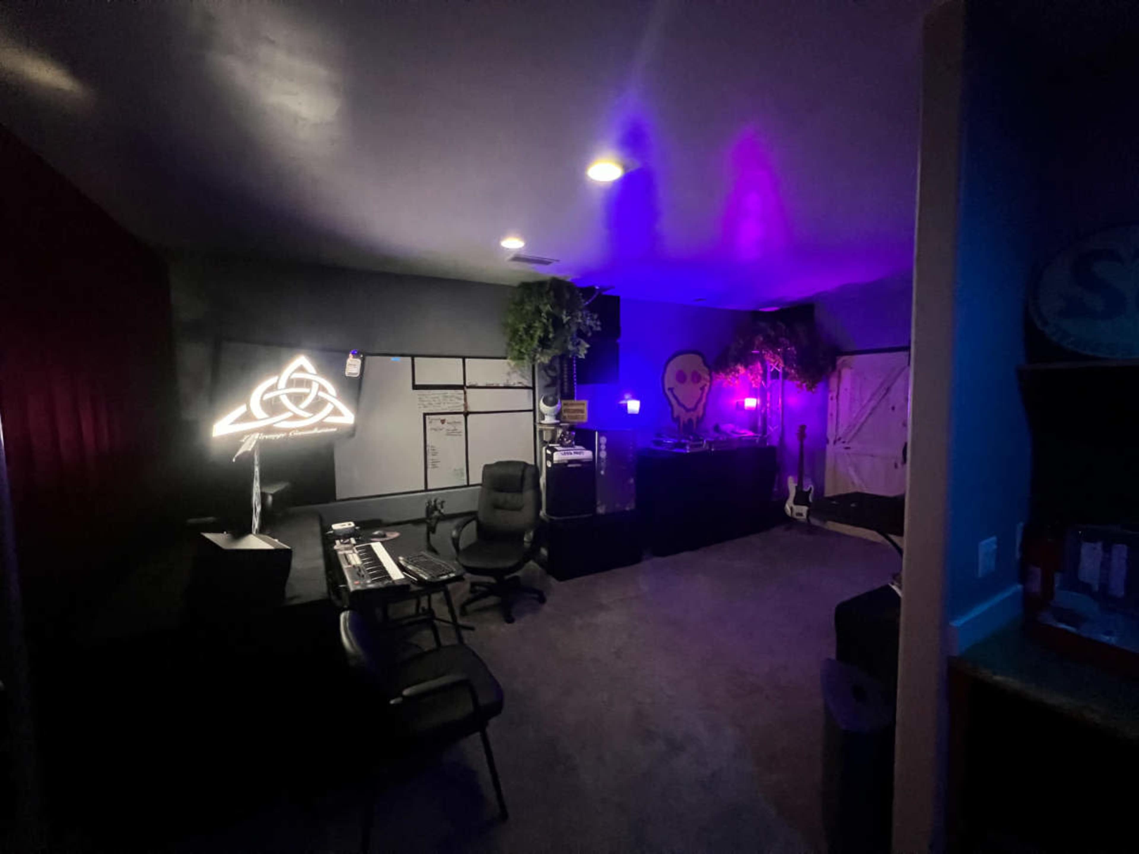 The image shows a dimly lit music studio featuring a keyboard, chairs, colorful neon lights, and musical instruments against a dark wall.