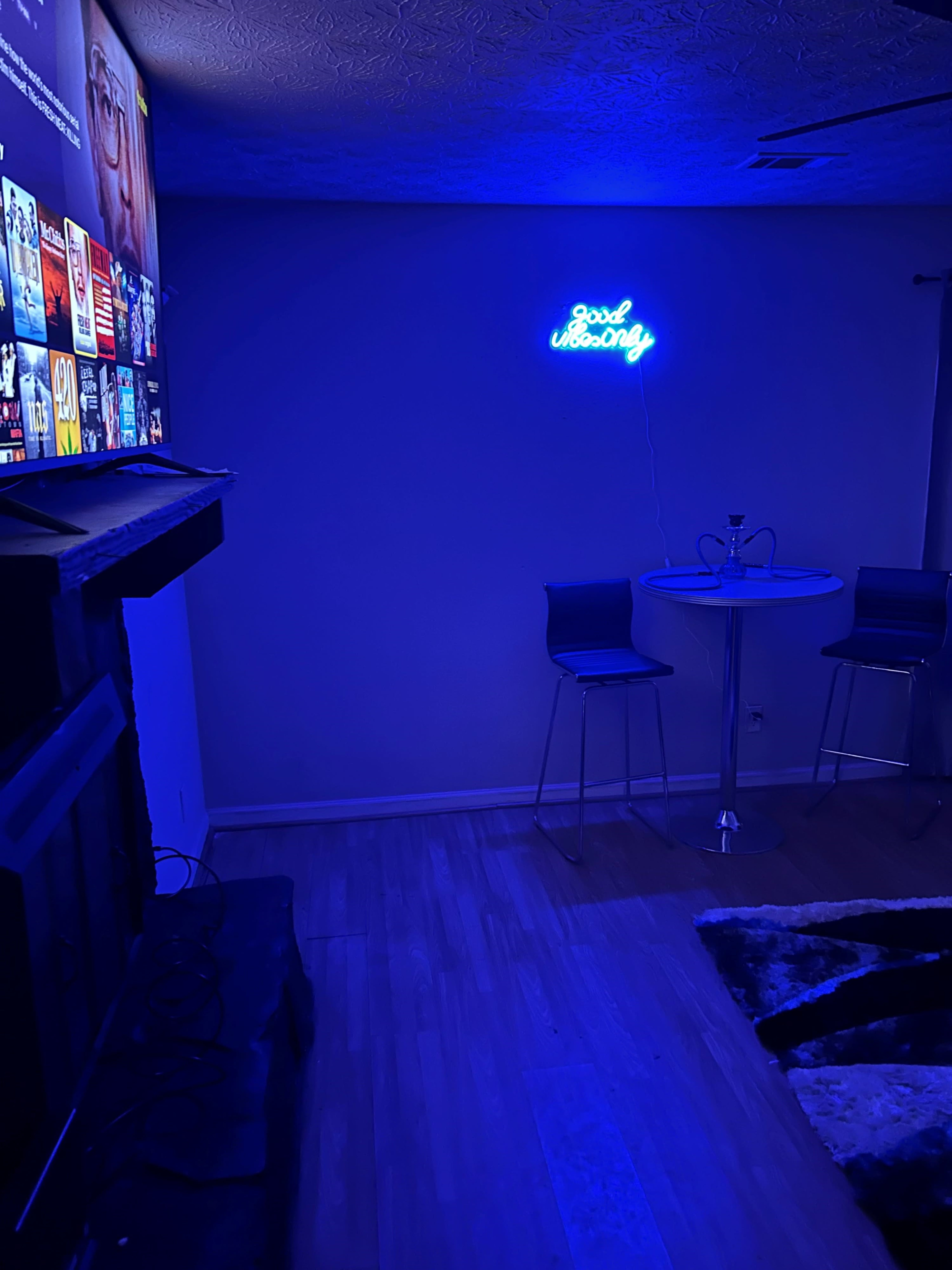 A dimly lit living room with a glowing blue neon sign that says "Good Morning," a small round table, and two metal chairs.