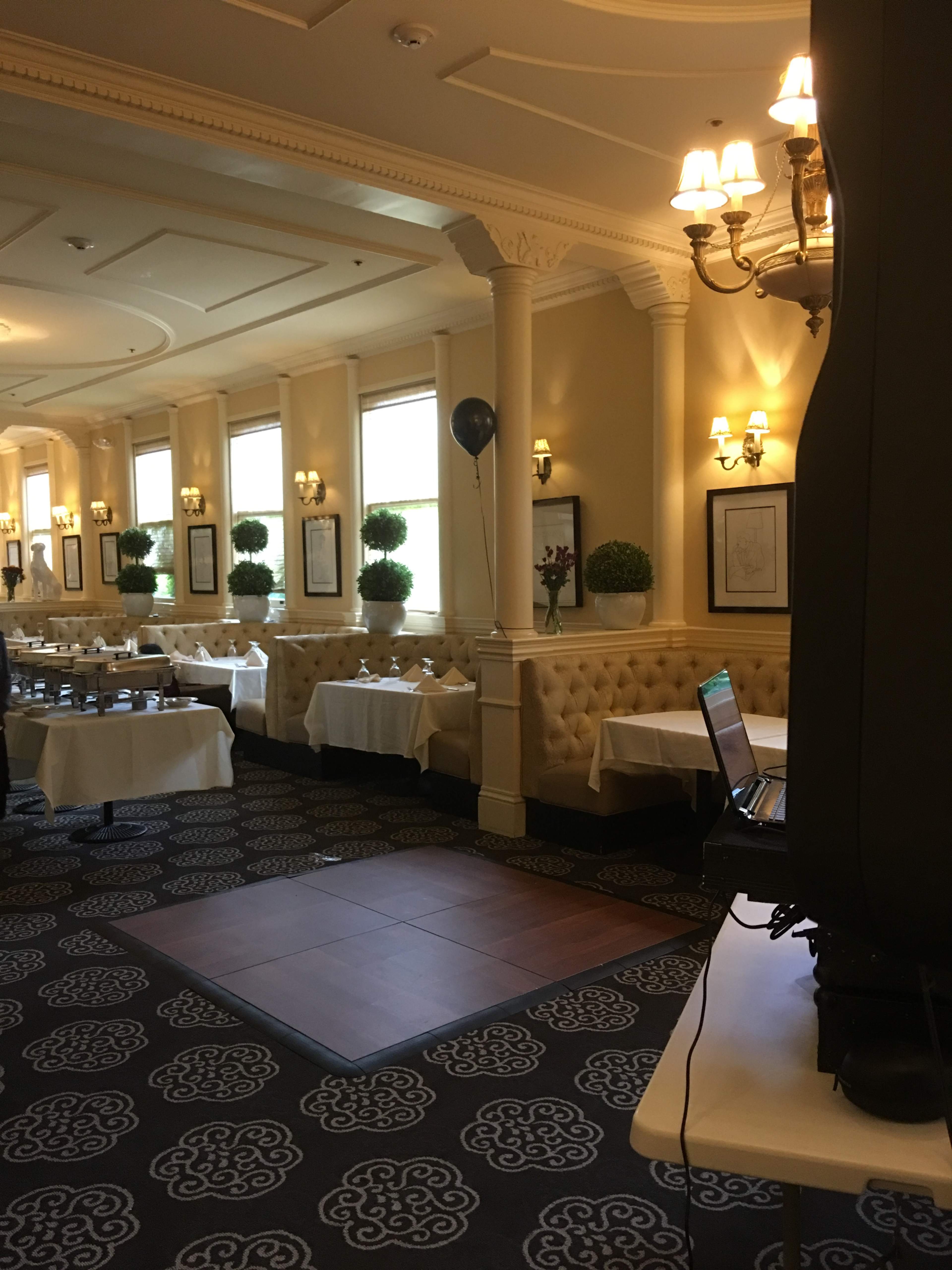 A well-lit restaurant interior with neatly arranged tables, upholstered seating, and decorative plants along the walls.