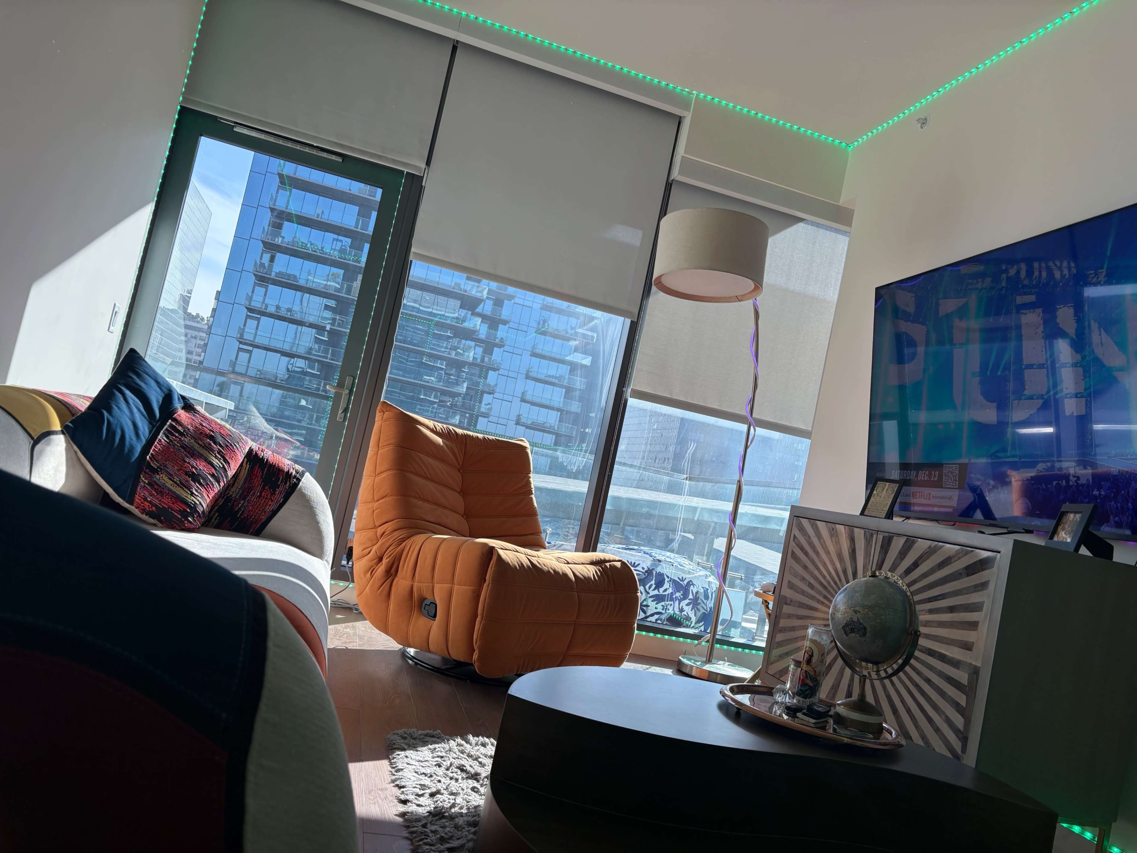 The image shows a modern living room with a bright orange chair, a television, and large windows allowing natural light to fill the space.