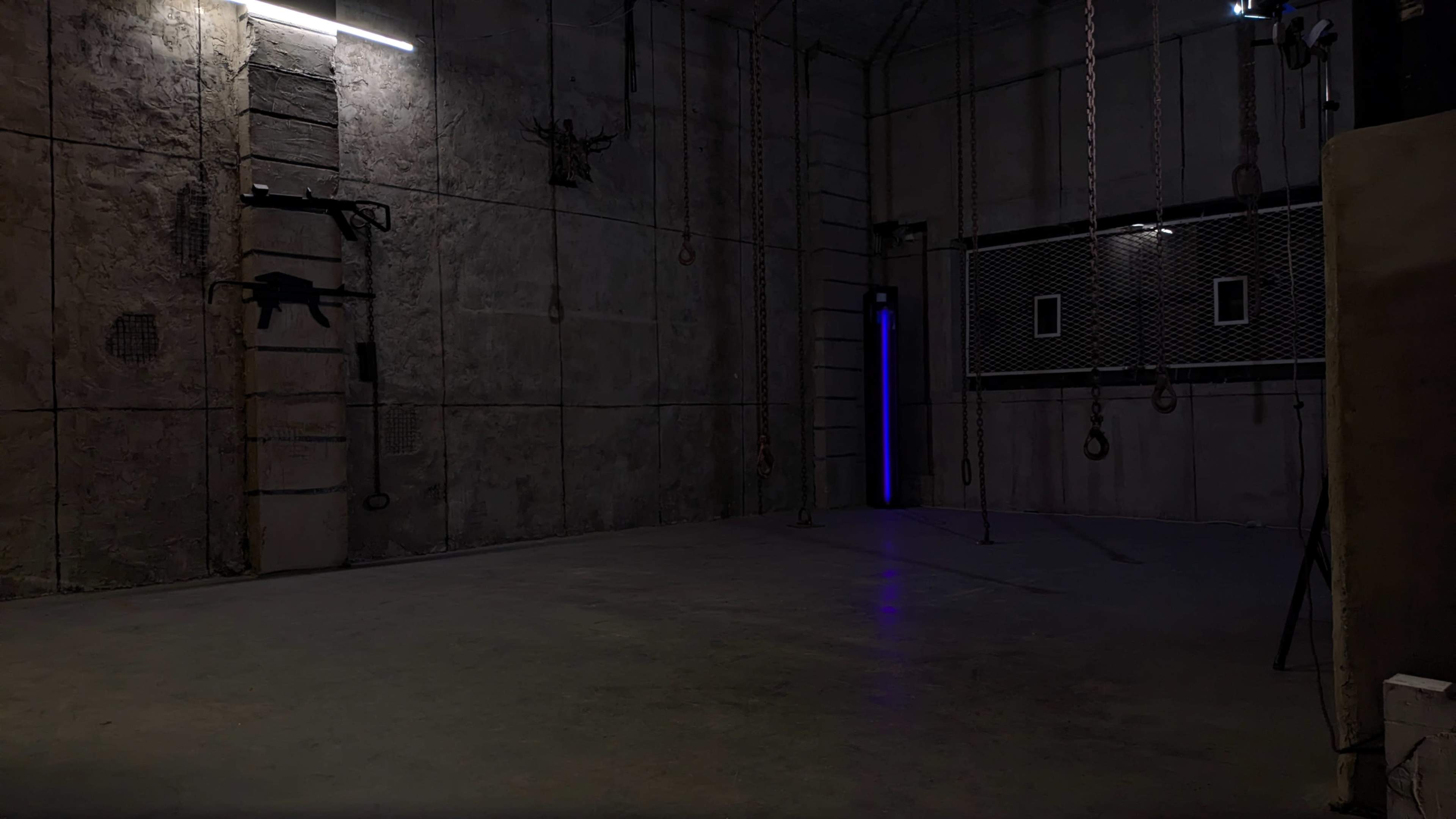 The image shows a dimly lit industrial room with concrete walls, featuring various hanging ropes and chains, and a single blue light illuminating one corner.