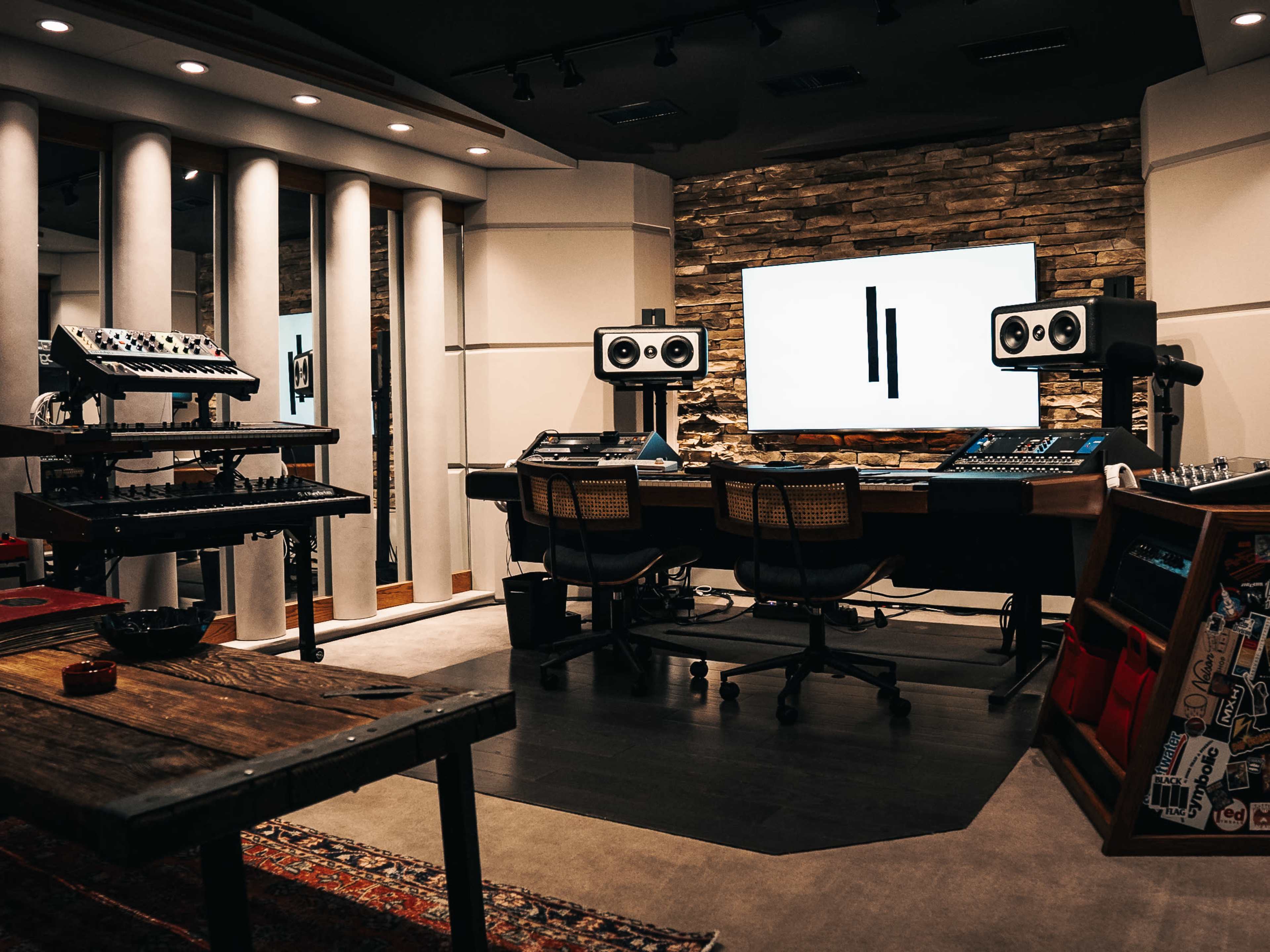 The image shows a modern music production studio featuring a large mixing desk, studio monitors, and various electronic instruments against a backdrop of wooden paneling and softly-lit walls.