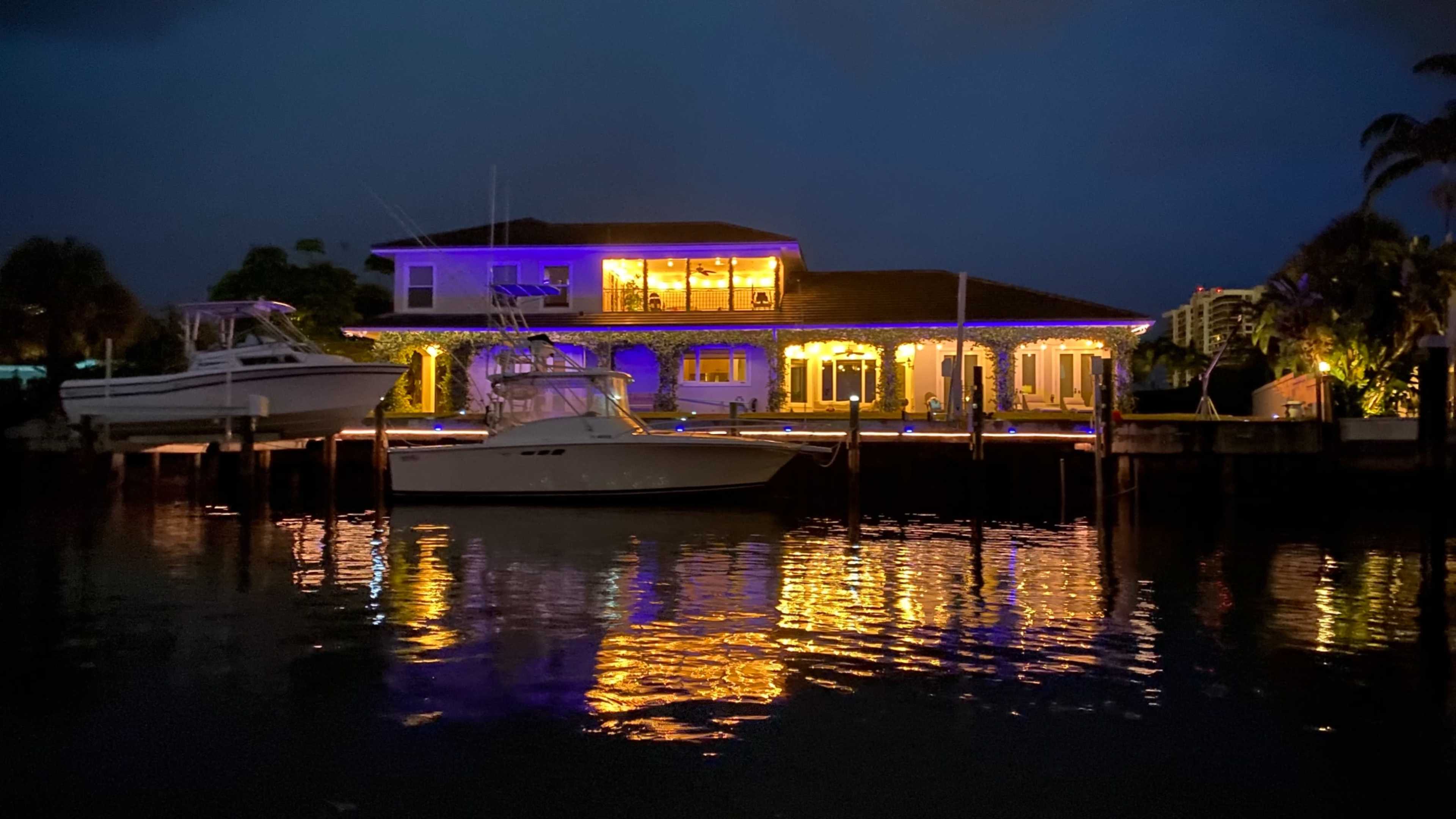 A waterfront house with colorful lights reflects on the water beside two moored boats.