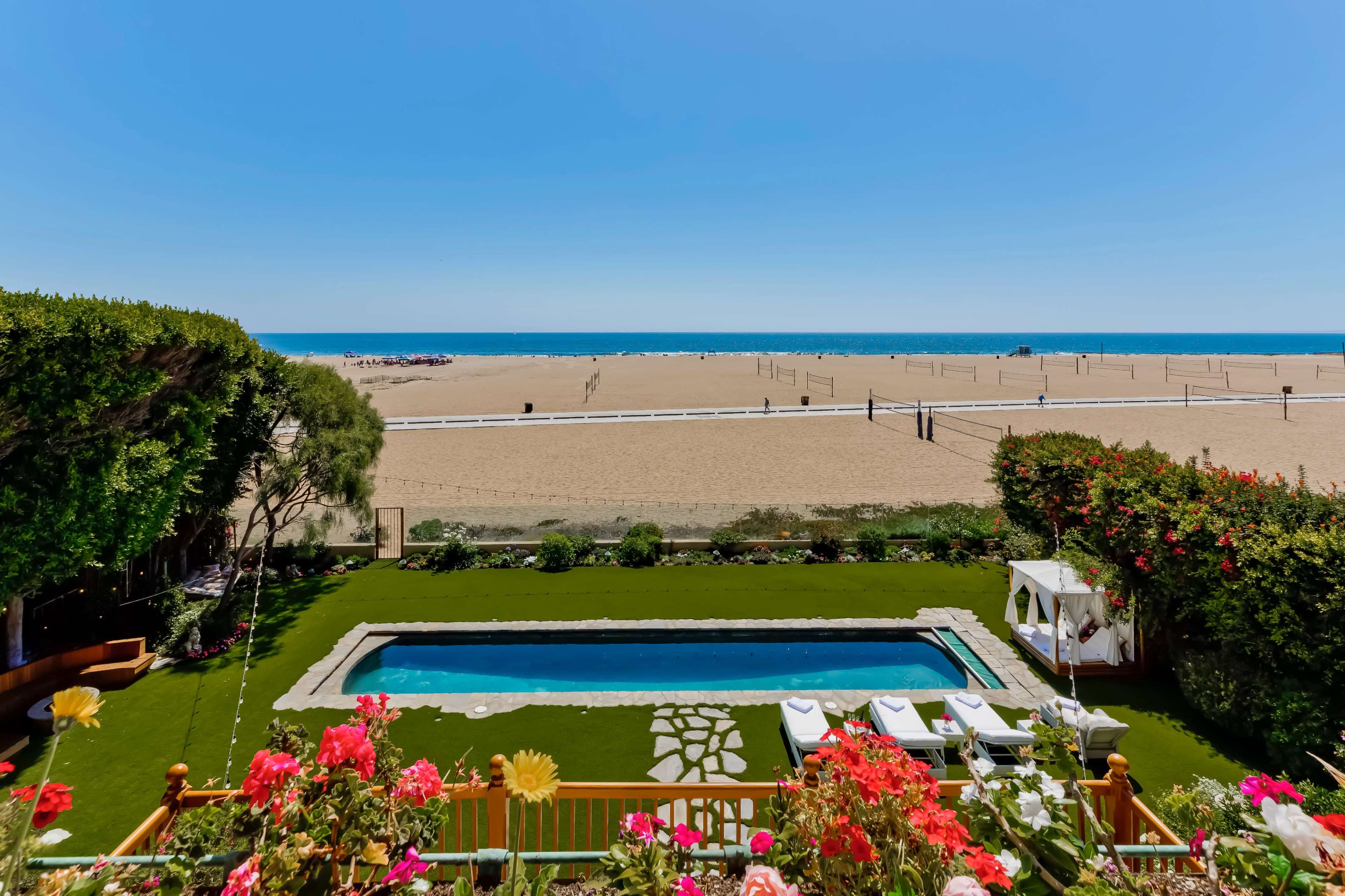 The image shows a backyard with a swimming pool surrounded by green grass and colorful flowers, overlooking a sandy beach and the ocean in the distance.