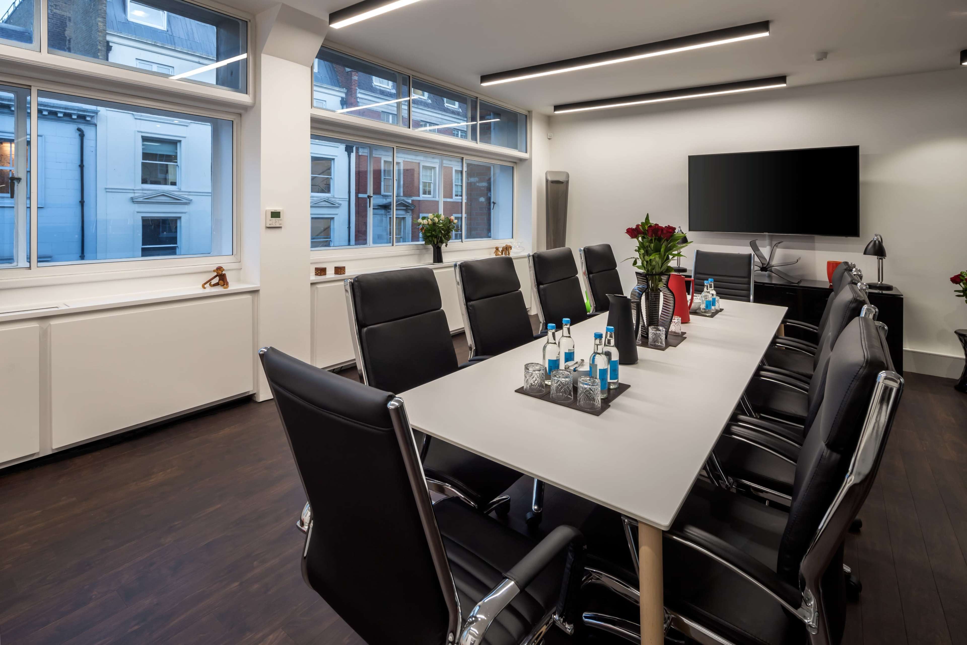 A modern conference room featuring a long table surrounded by black chairs, large windows with natural light, and a wall-mounted television.