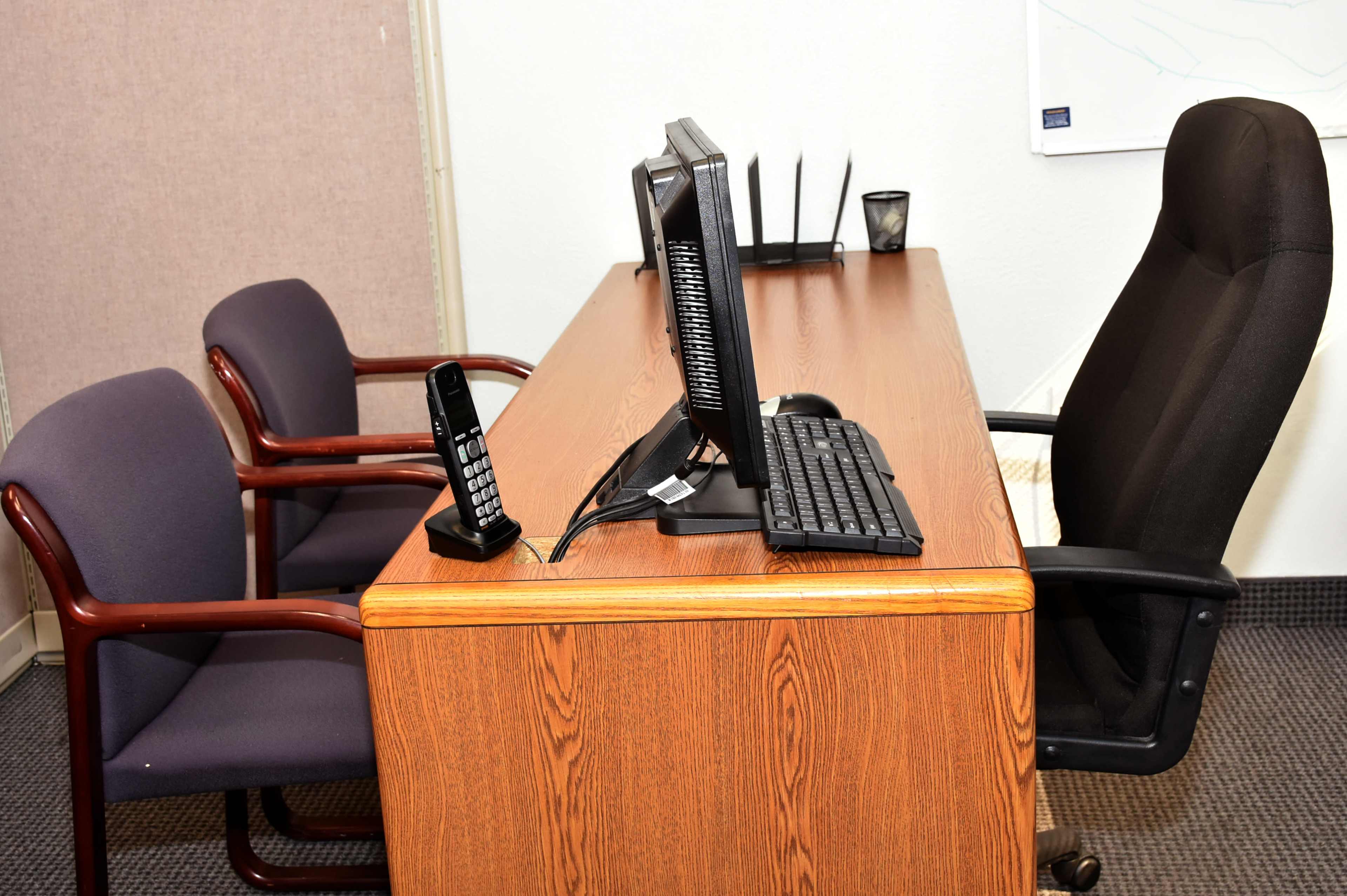 A desk with a computer monitor, keyboard, and an office phone, flanked by two chairs in a simple office space.