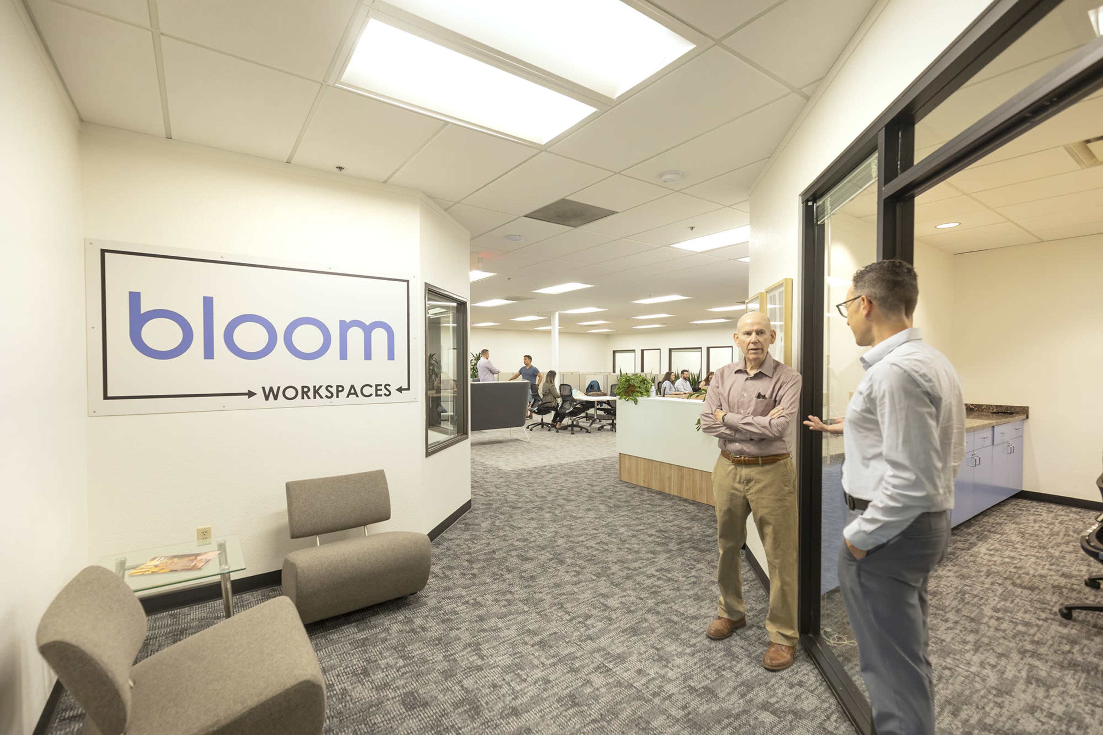 Two men stand conversing near the entrance of an office space featuring the logo "bloom workspaces" on the wall, with several people working at desks in the background.