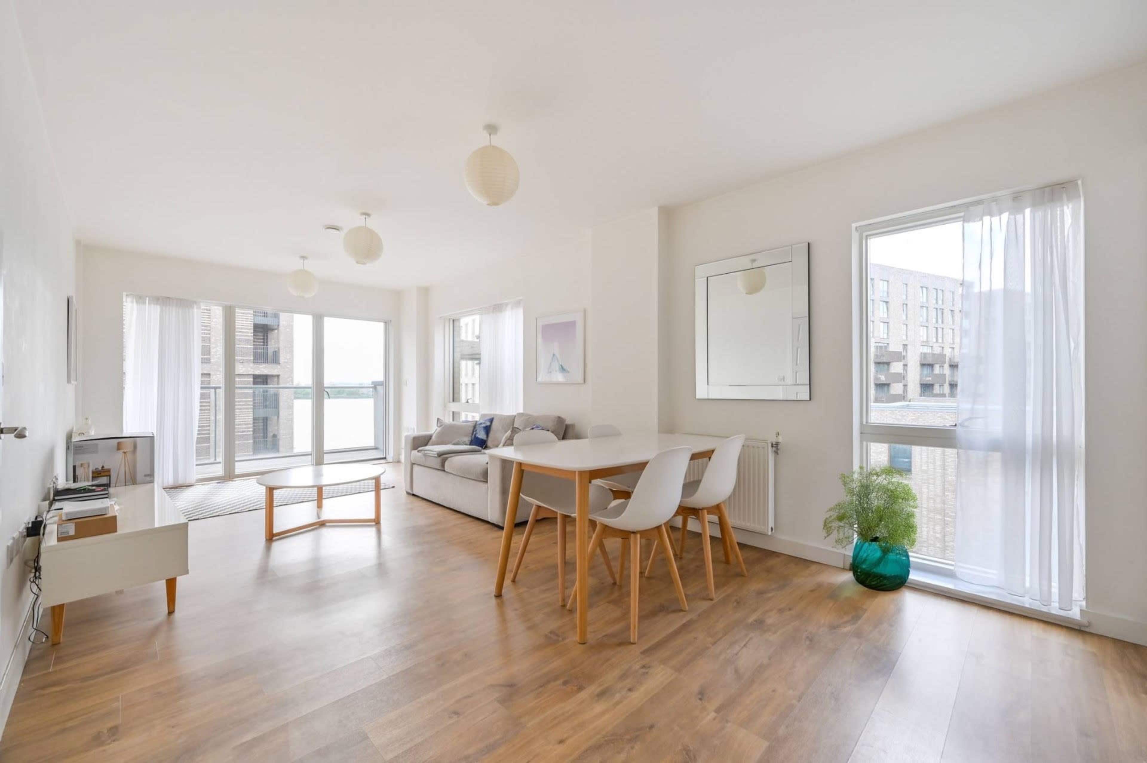 The image shows a bright, modern living room and dining area with large windows, light wood flooring, and minimalist furniture.