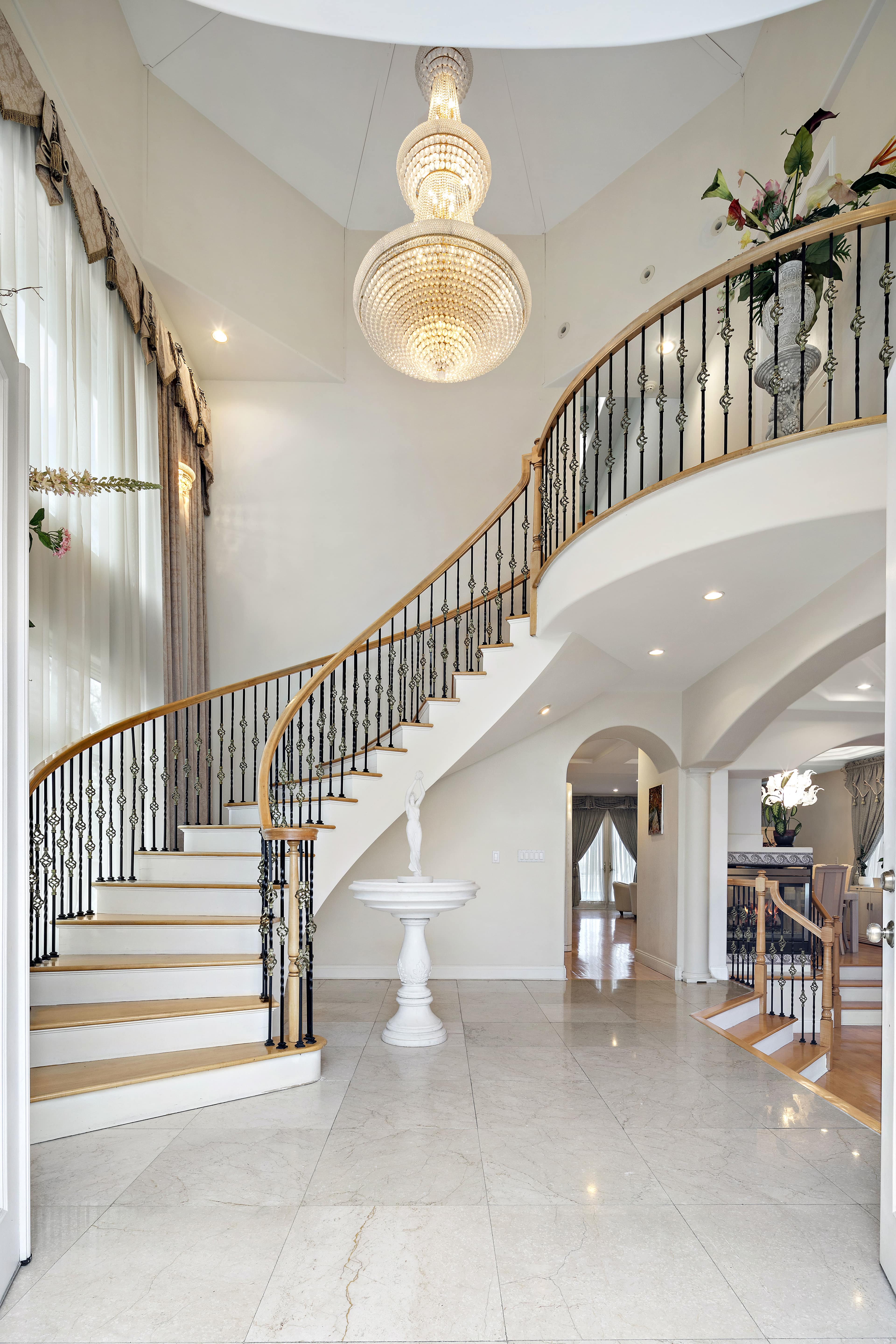 An elegant foyer features a curved staircase with a wooden banister, a large chandelier above, and marble flooring.