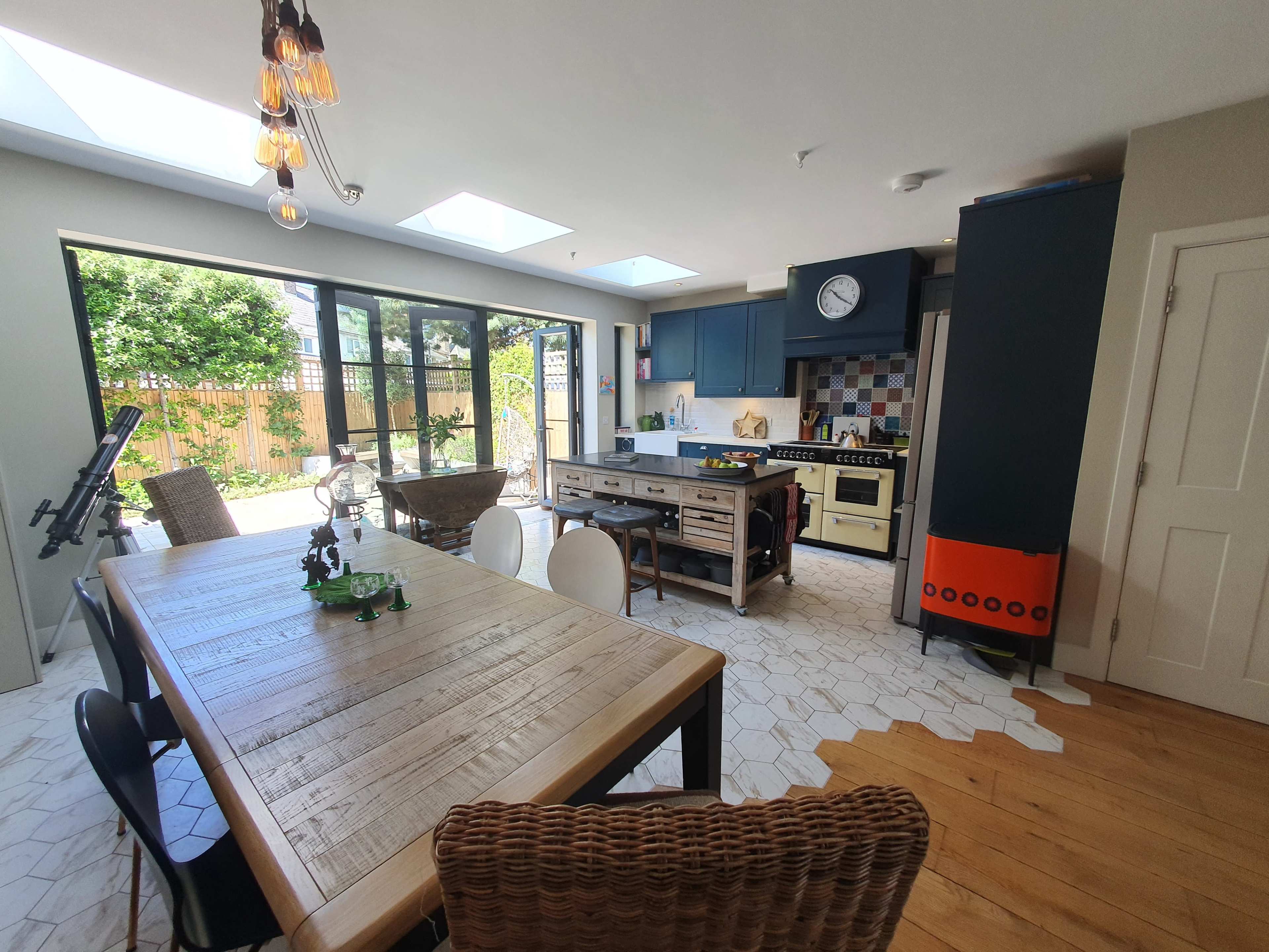 A modern kitchen and dining area with a wooden table, various appliances, and large windows that open to a garden.