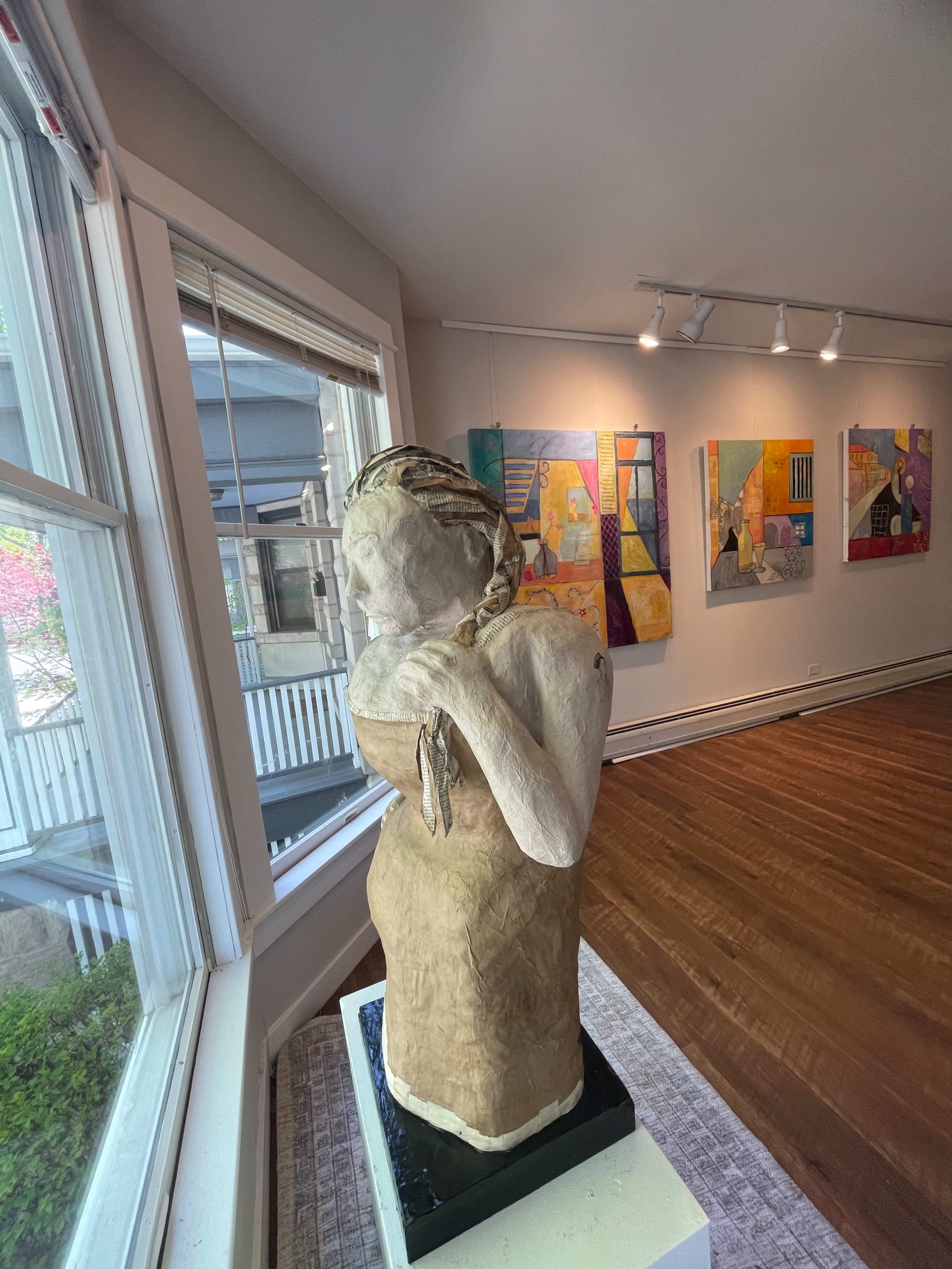 A statue of a woman with a towel wrapped around her body stands on a pedestal in a gallery filled with colorful abstract paintings.