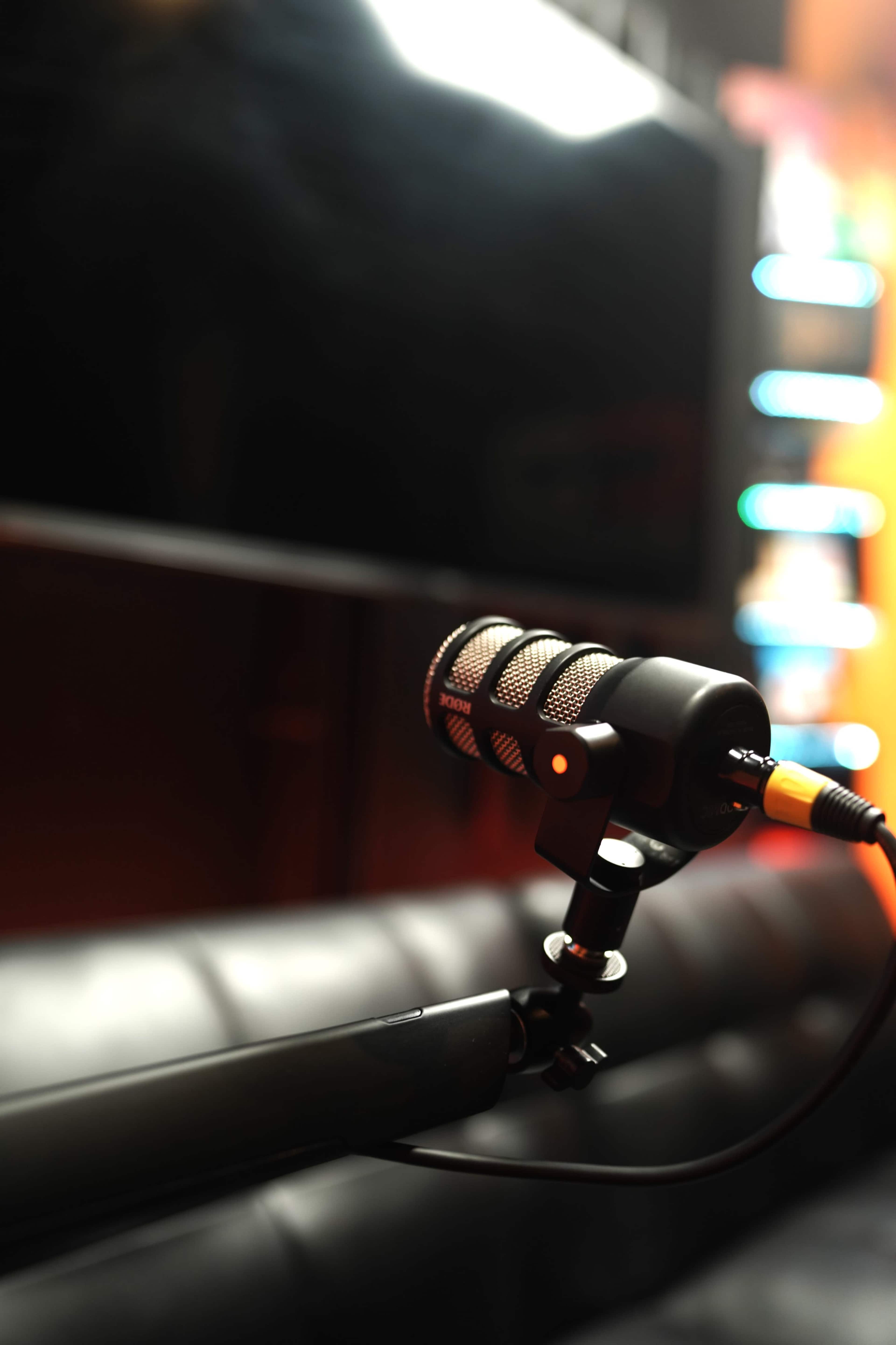 A close-up of a black microphone with a textured grille attached to a stand, positioned in front of a blurred background featuring a screen and soft lighting.
