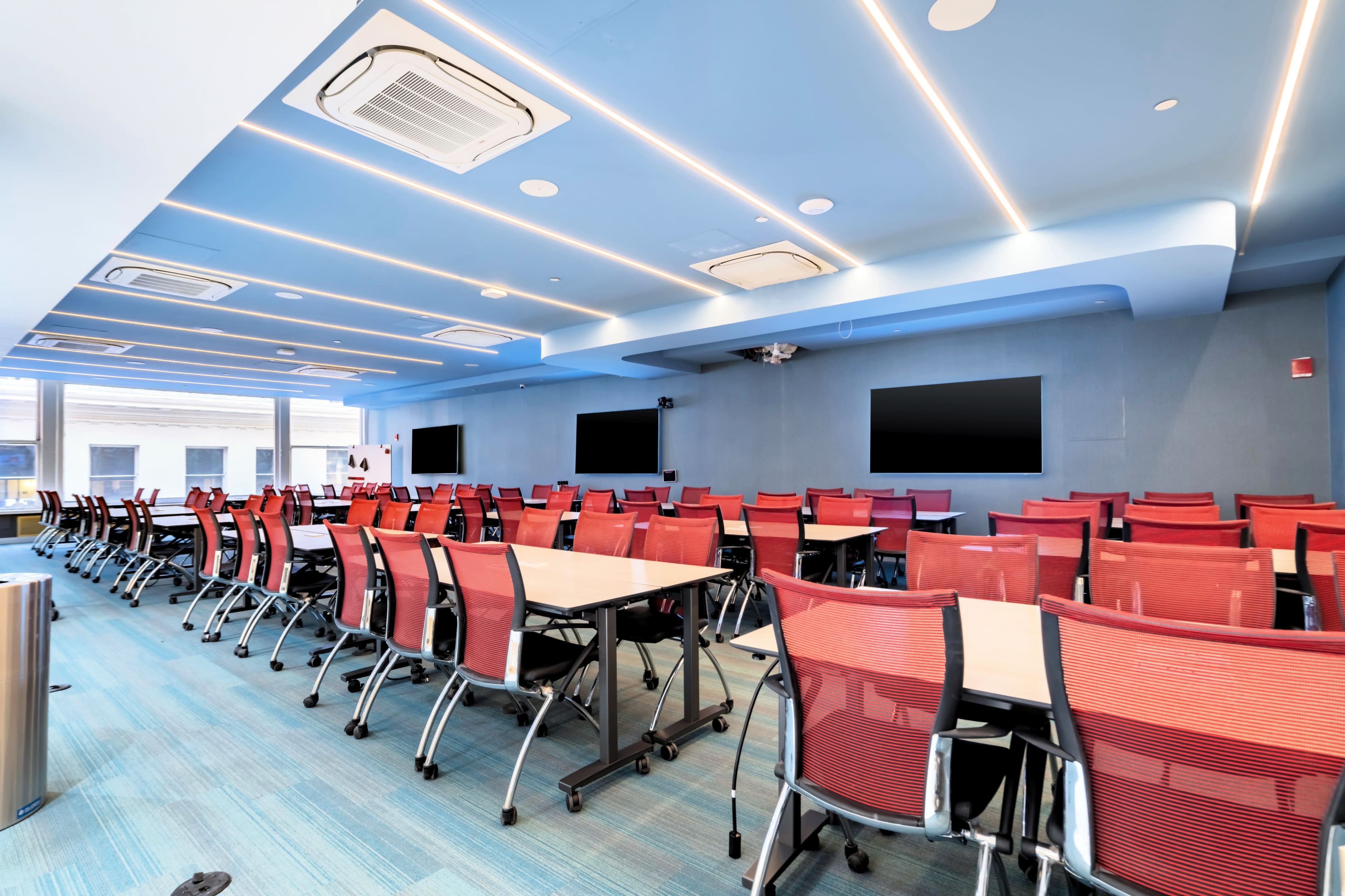 The image shows a modern conference room with rows of red mesh chairs and large flat-screen monitors on the walls.