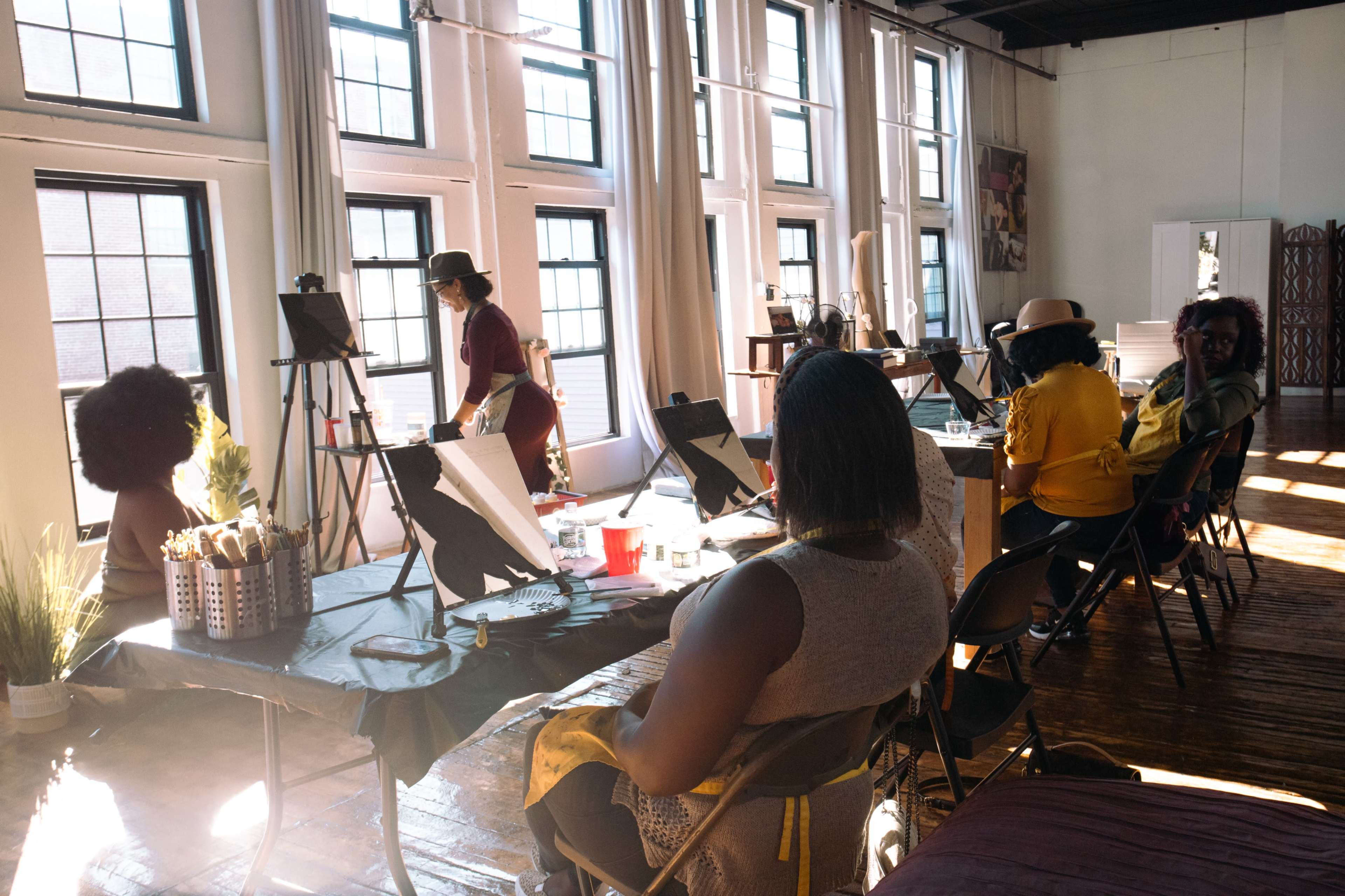 A group of individuals is engaged in an art class, seated at tables with easels in a sunlit studio featuring large windows.