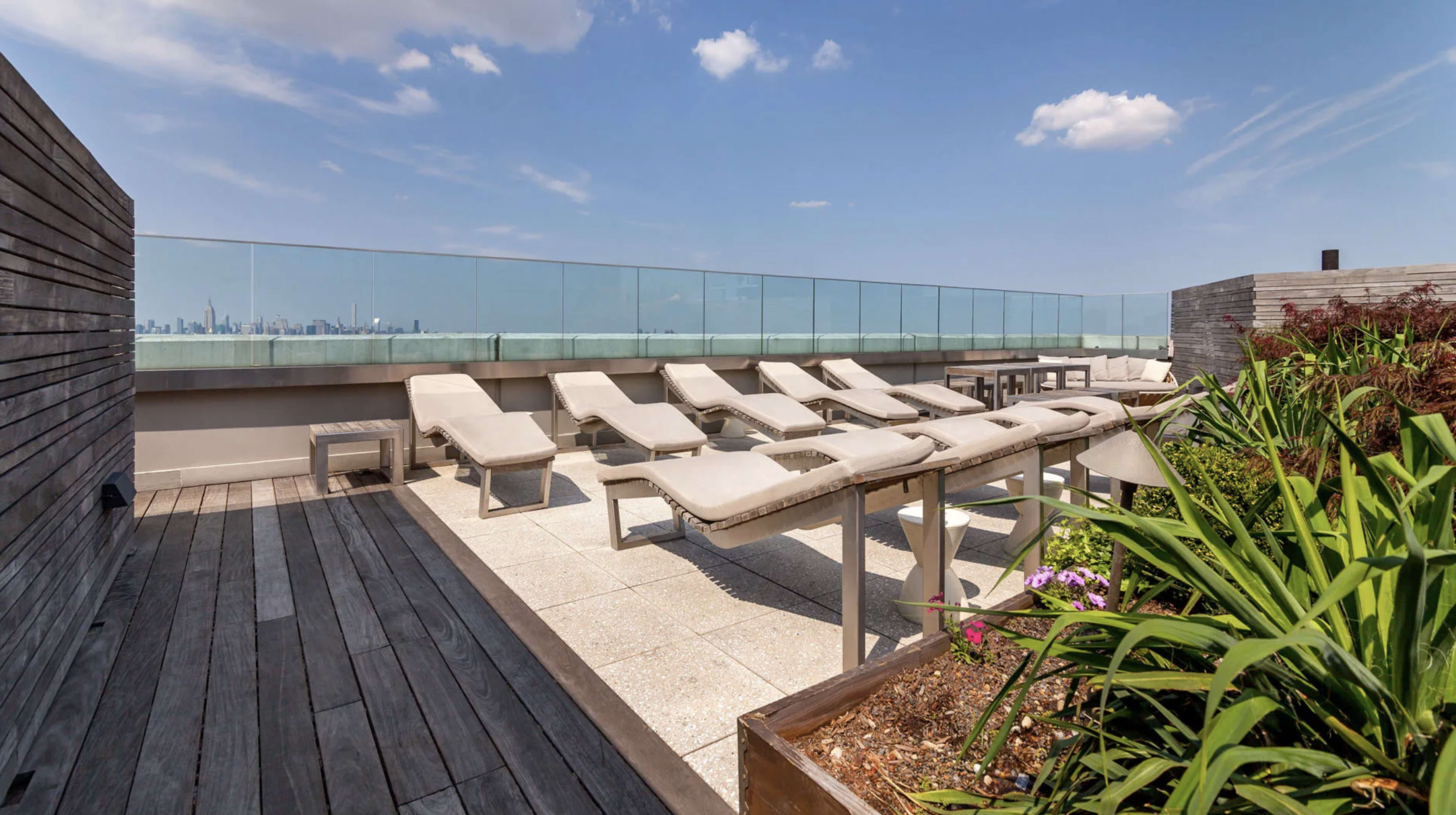 The image shows a rooftop terrace featuring several lounge chairs, a glass railing, and planters with greenery.