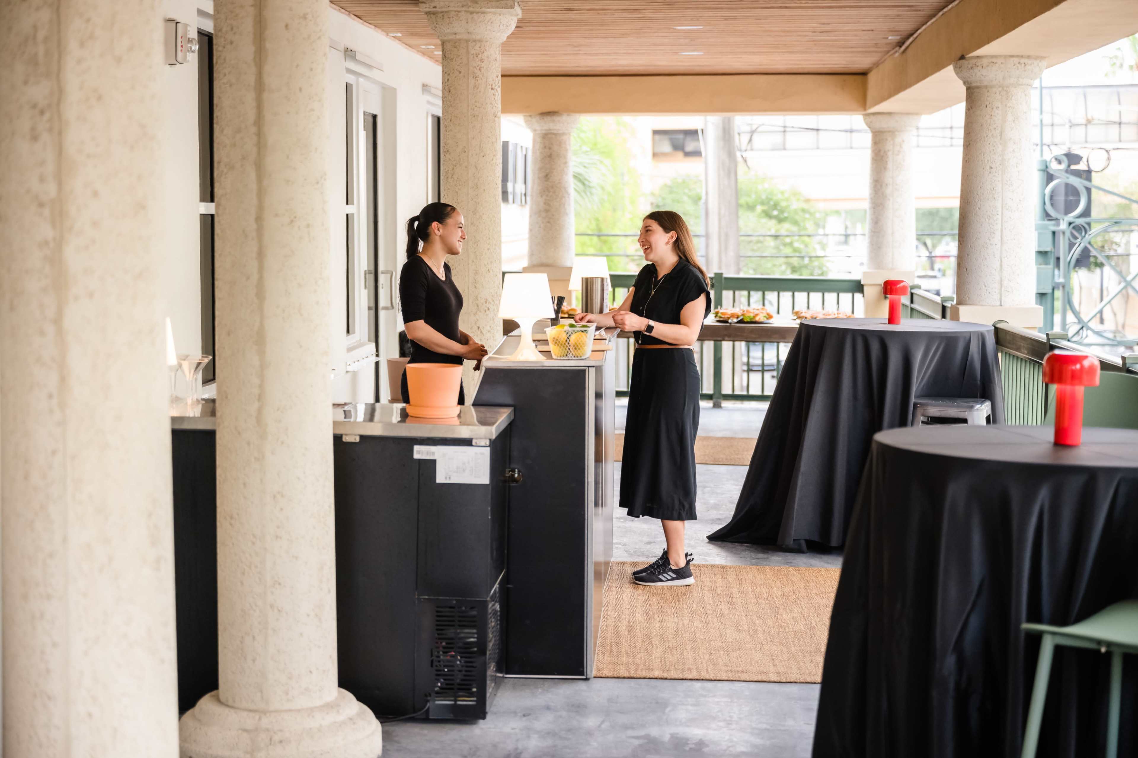 Two women converse at a coffee service station with black tables and food set up in the background.