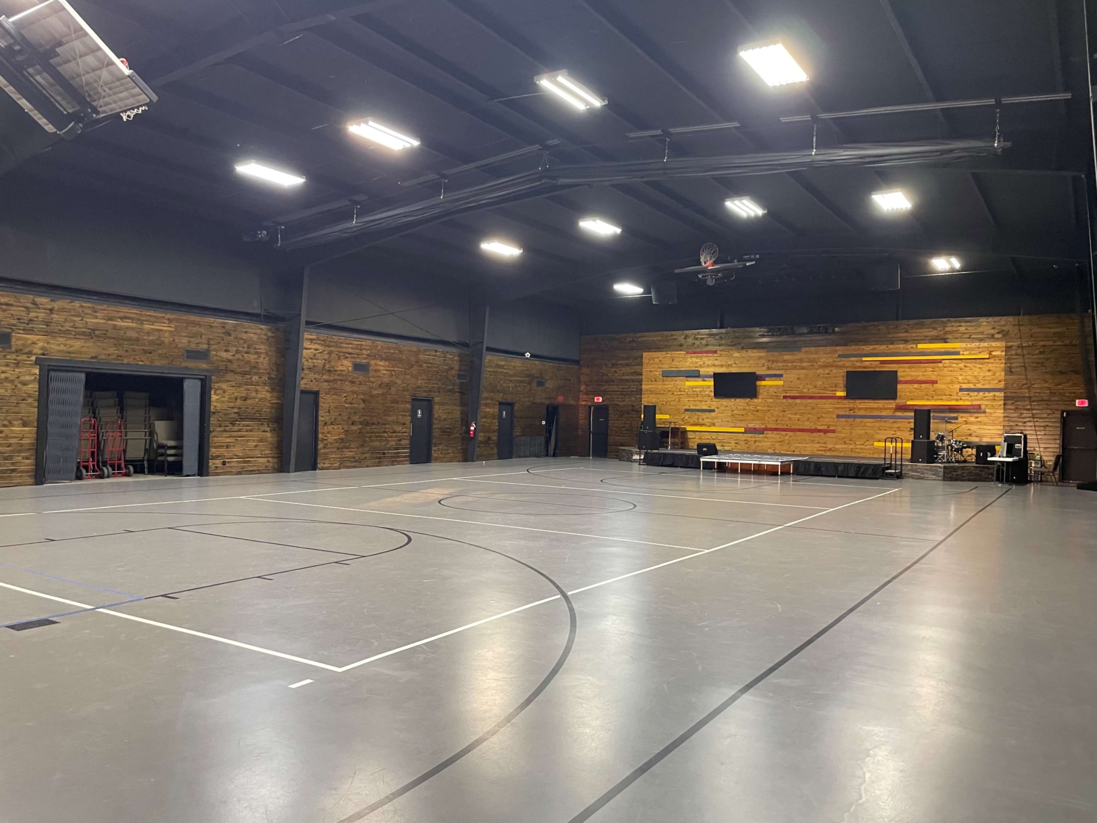 The image showcases a large indoor gymnasium with a polished gray floor, wooden accent walls, and various sports equipment arranged around the space.