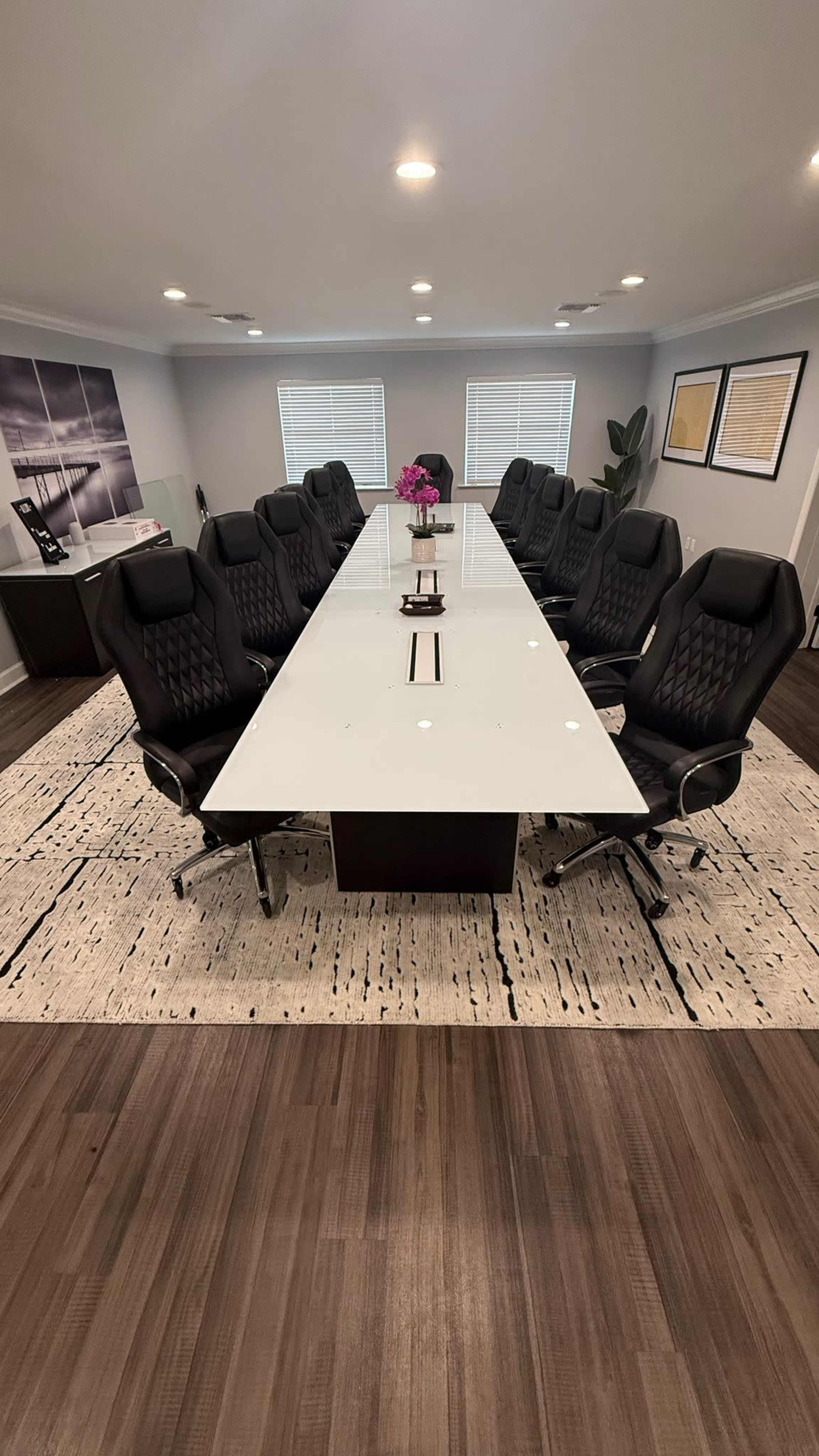 A modern conference room features a long white table surrounded by ergonomic black chairs on a patterned rug.