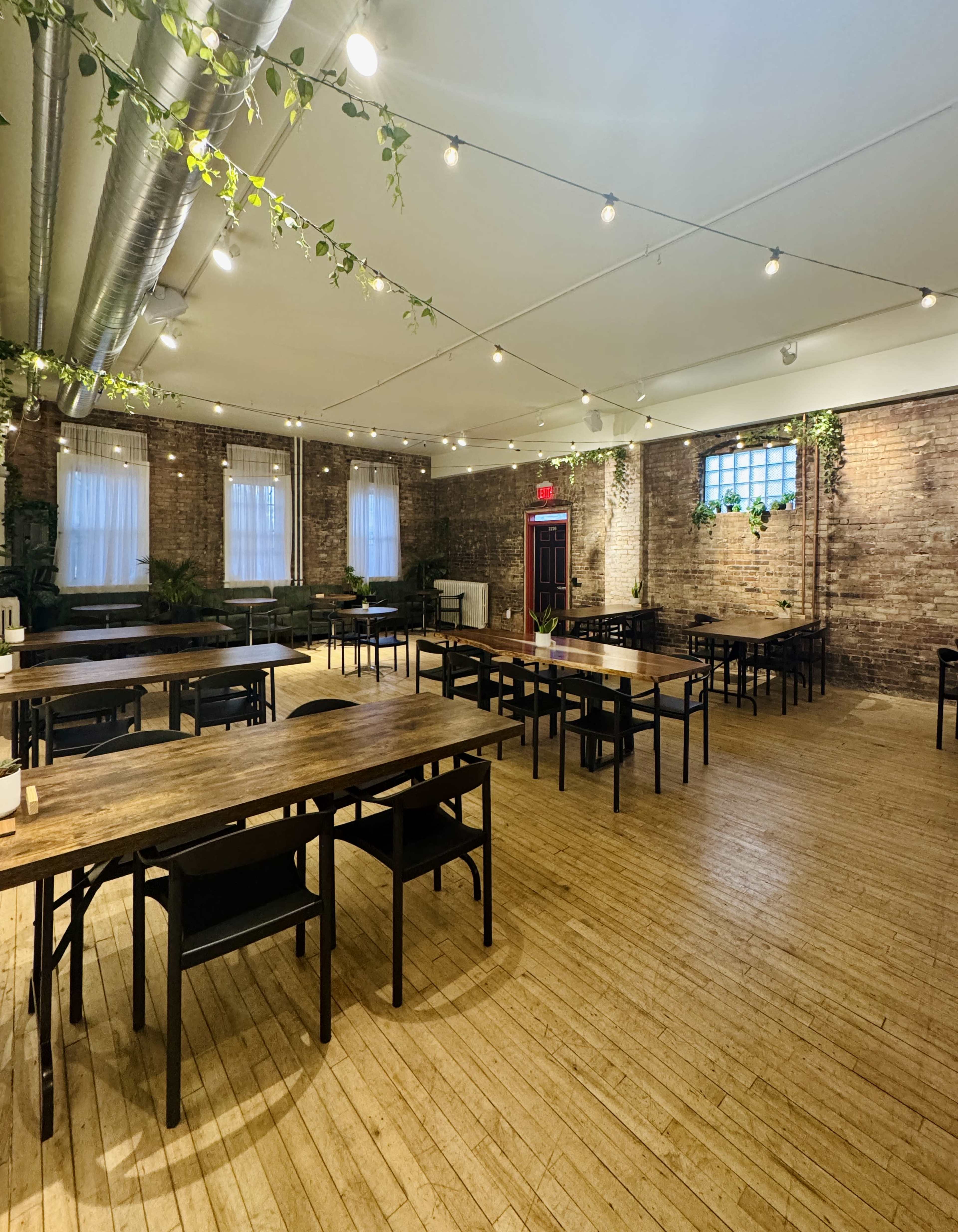 The image shows a spacious room with wooden tables and chairs arranged neatly, exposed brick walls, and string lights overhead, creating a well-lit environment.