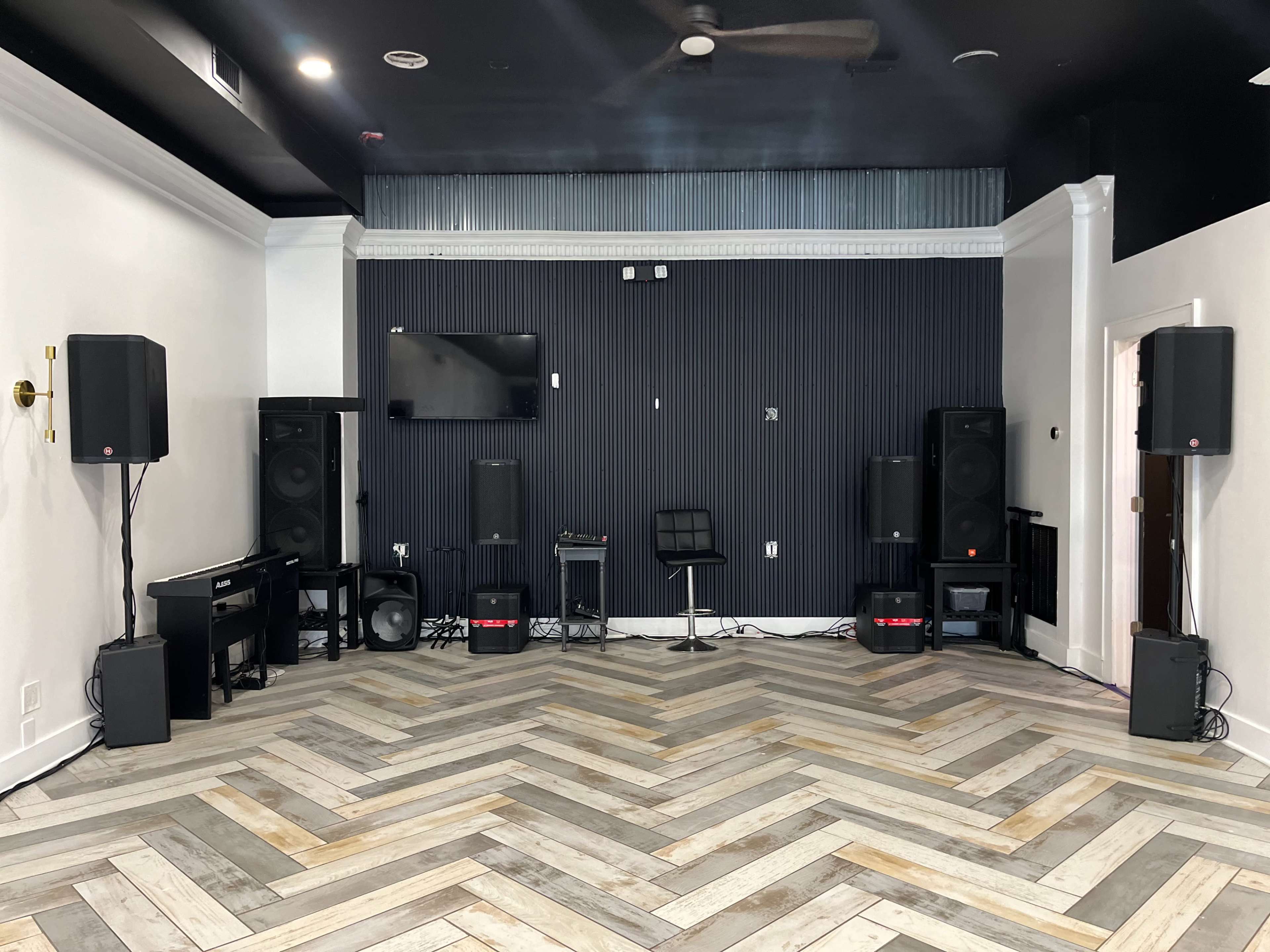 A sparsely furnished room with a herringbone wood floor, featuring speaker stacks, a television mounted on the wall, and a black chair positioned in the center.