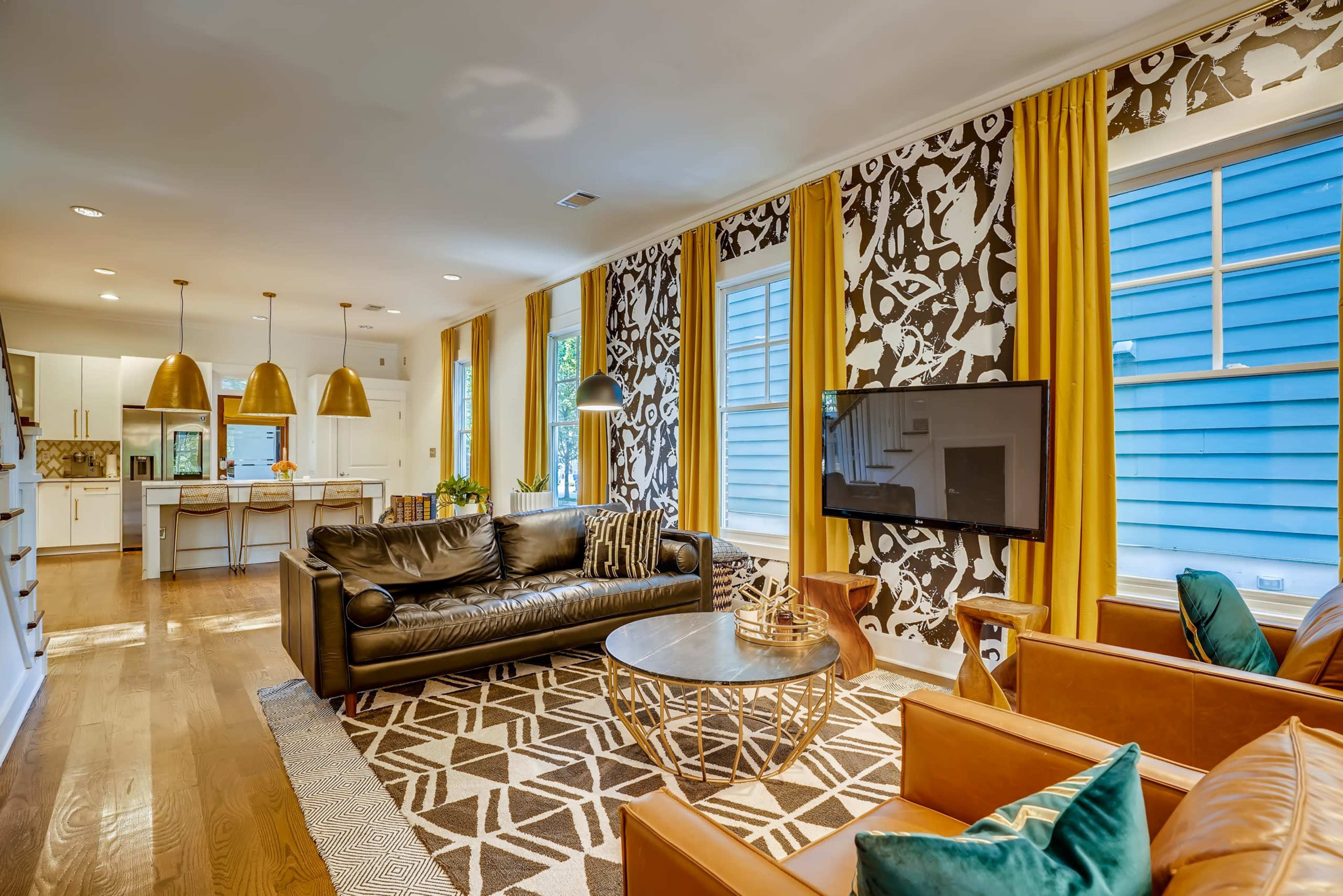The image shows a modern living room featuring a leather sofa, two brown armchairs, a round coffee table, and a patterned accent wall, with large windows and yellow curtains.