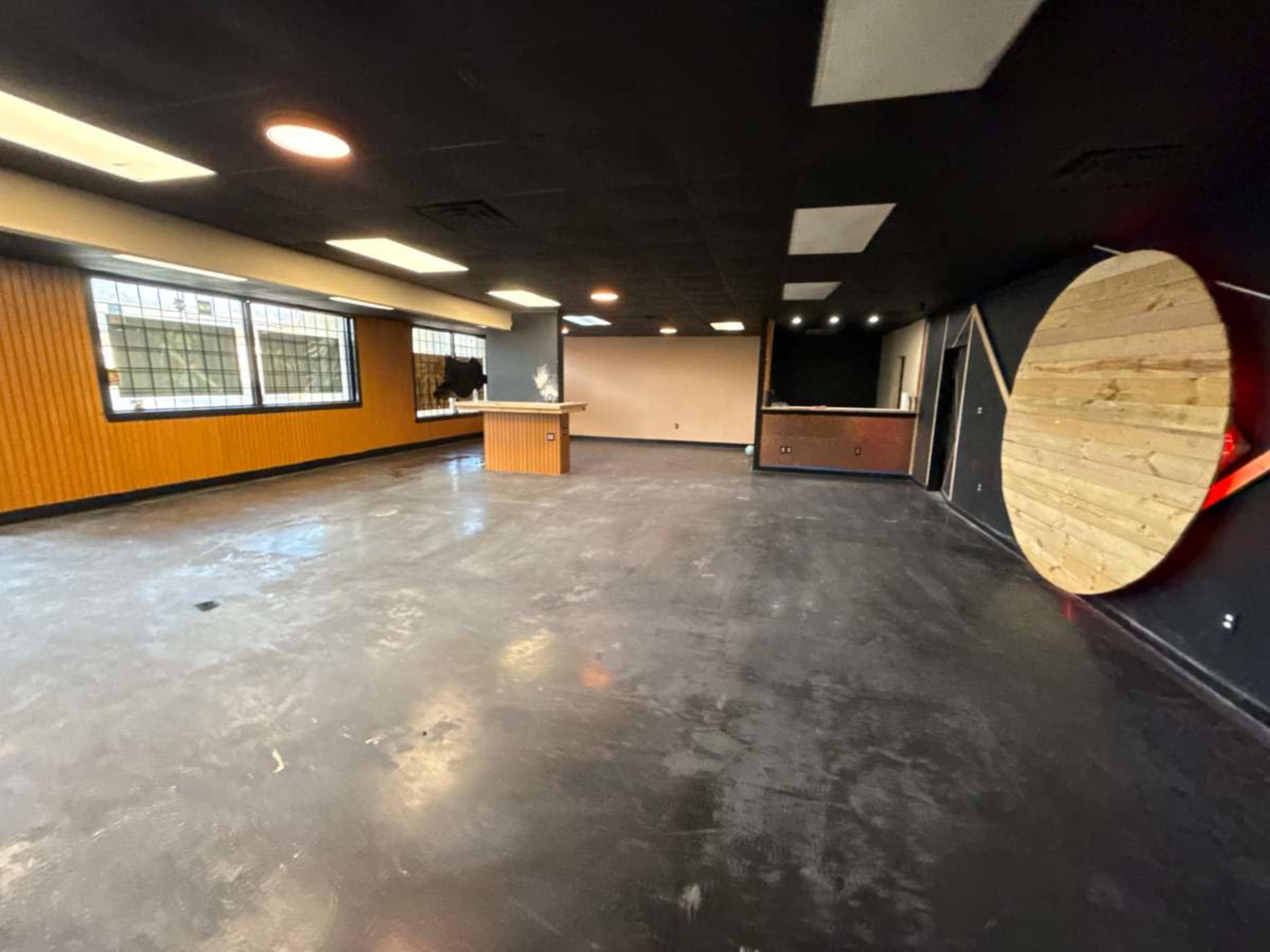 The image shows an empty room with a polished concrete floor, wooden accent walls, and a counter against one wall, along with a large circular wooden feature on another wall.