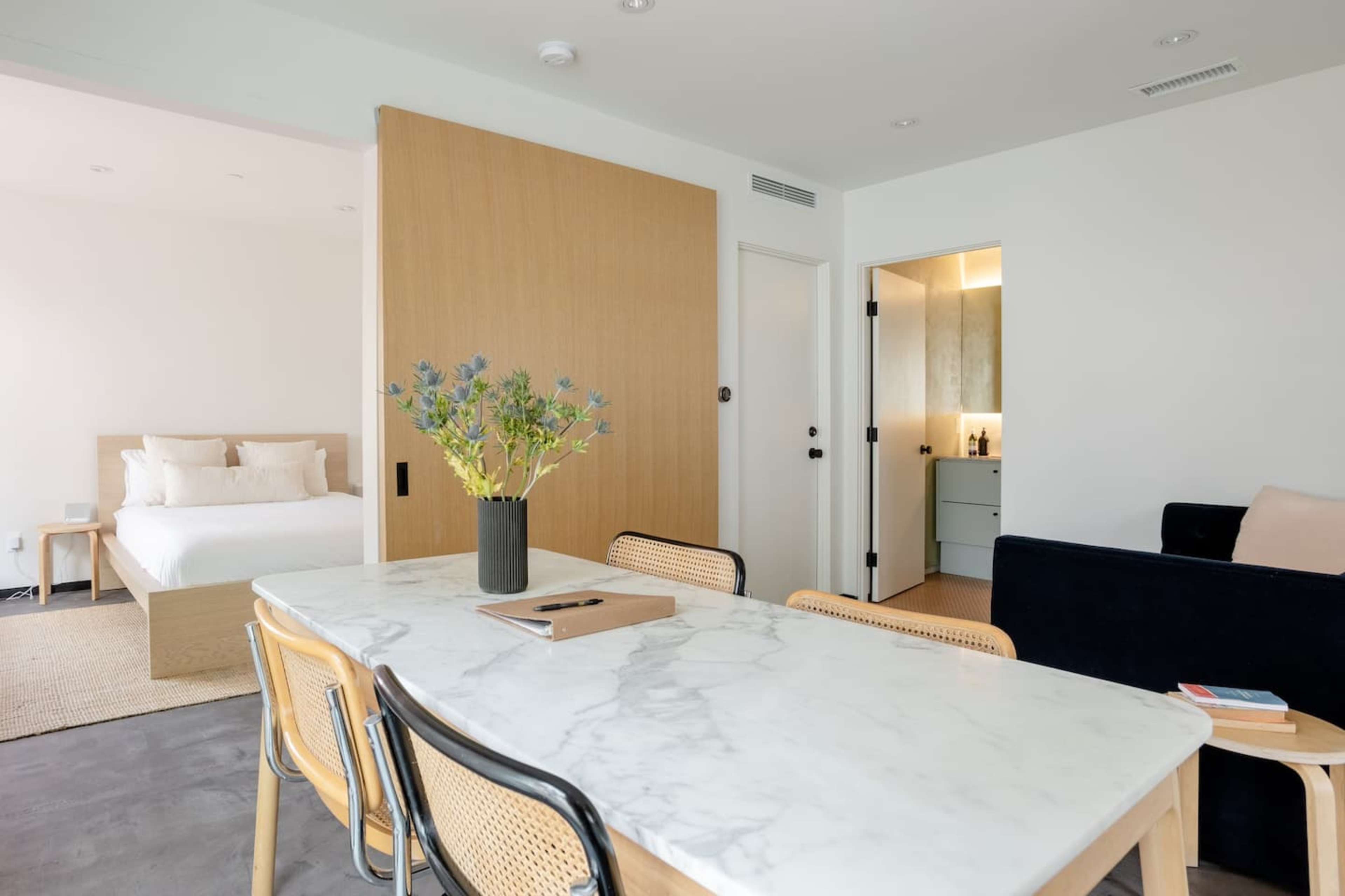 A modern living space features a marble dining table with wooden chairs, a bedroom with a bed visible through a sliding door, and a bathroom entrance in the background.
