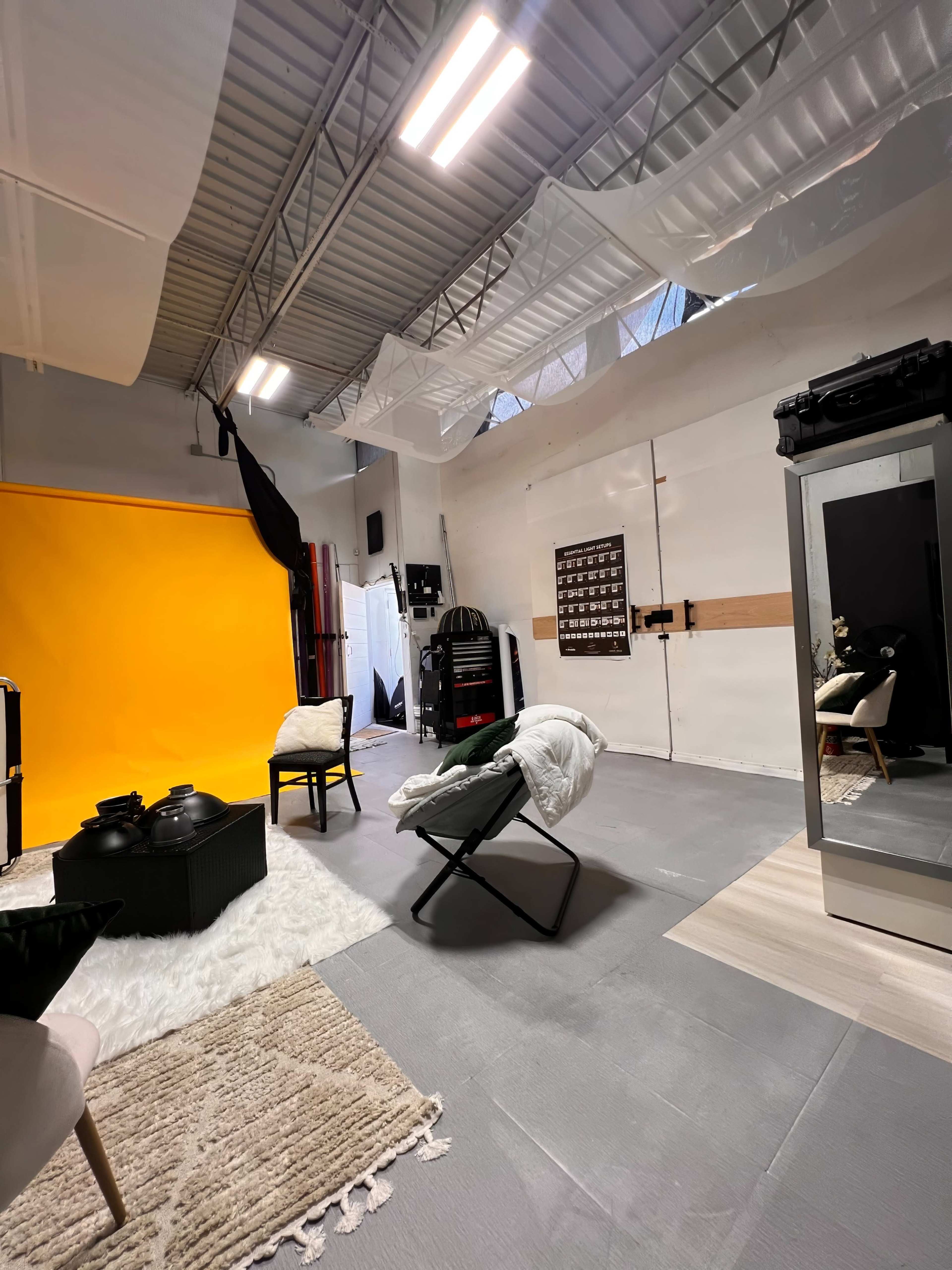 A spacious studio with a bright yellow backdrop, several chairs, a rug, and various photography equipment.