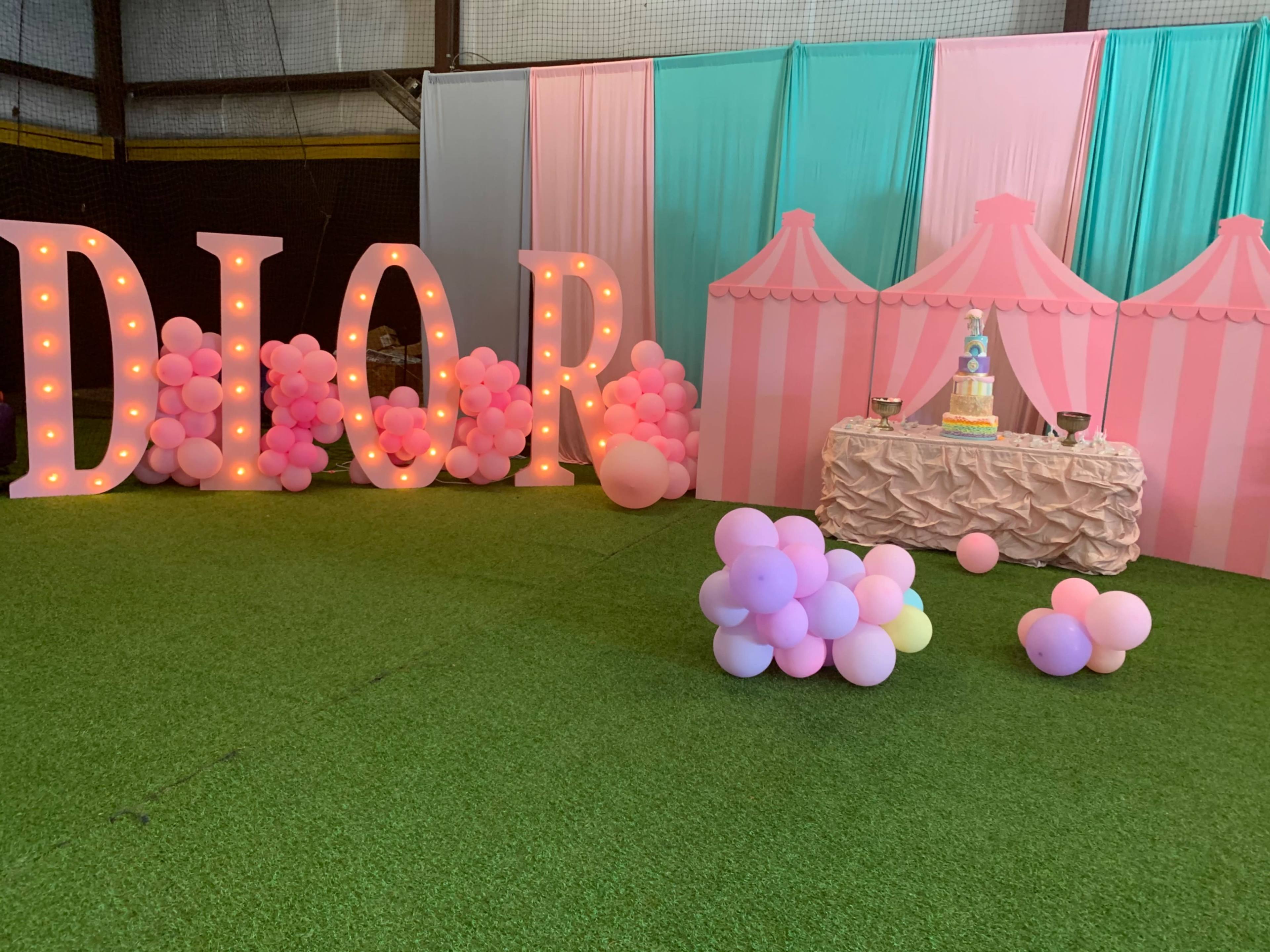 The scene features large illuminated "DIOR" letters and a decorative display with pink and blue striped tents, complemented by a table showcasing a cake and colorful balloons on the ground.