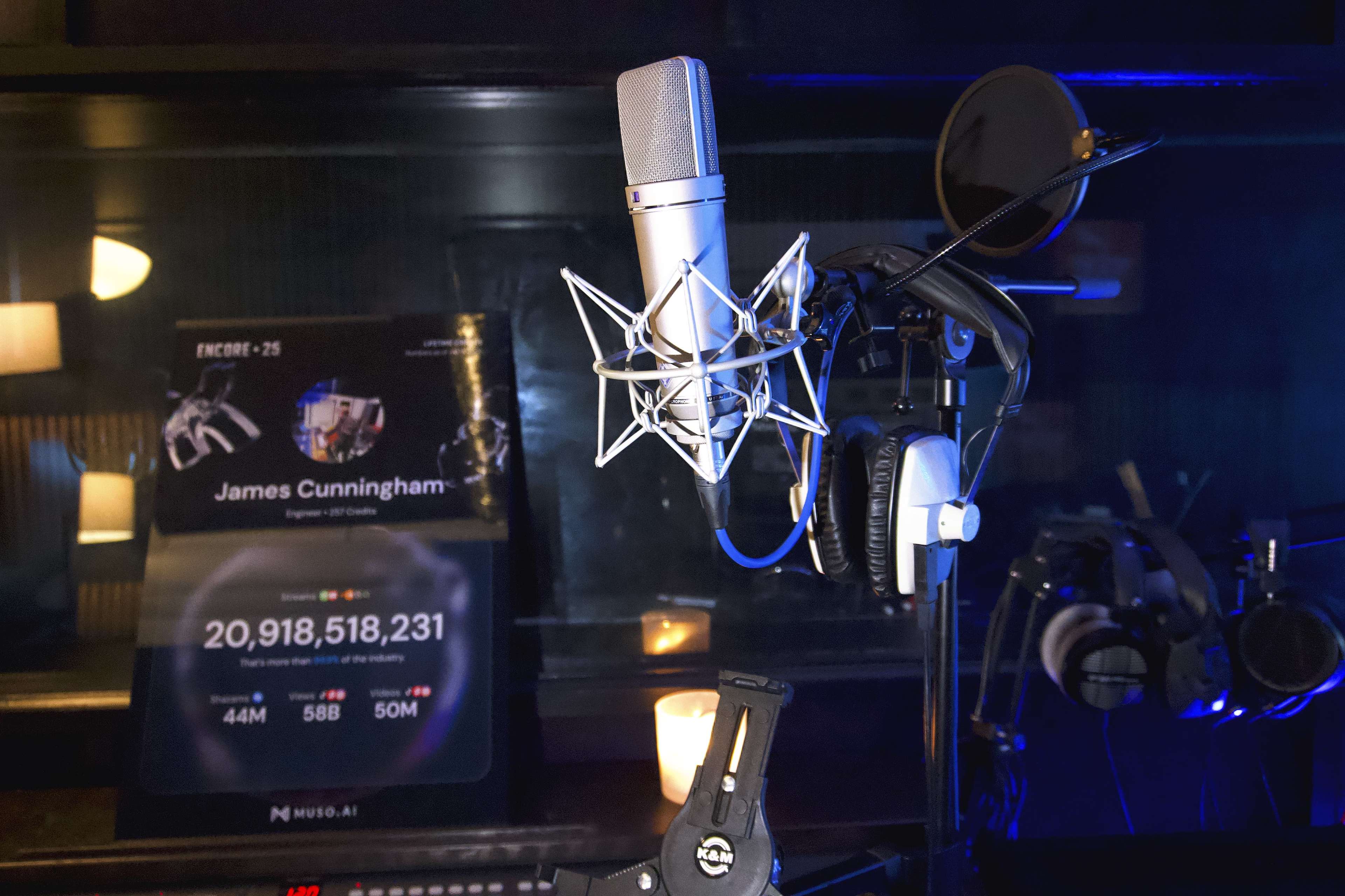A microphone is positioned on a stand in a dimly lit studio, with headphones hanging nearby and a digital display showcasing statistics in the background.