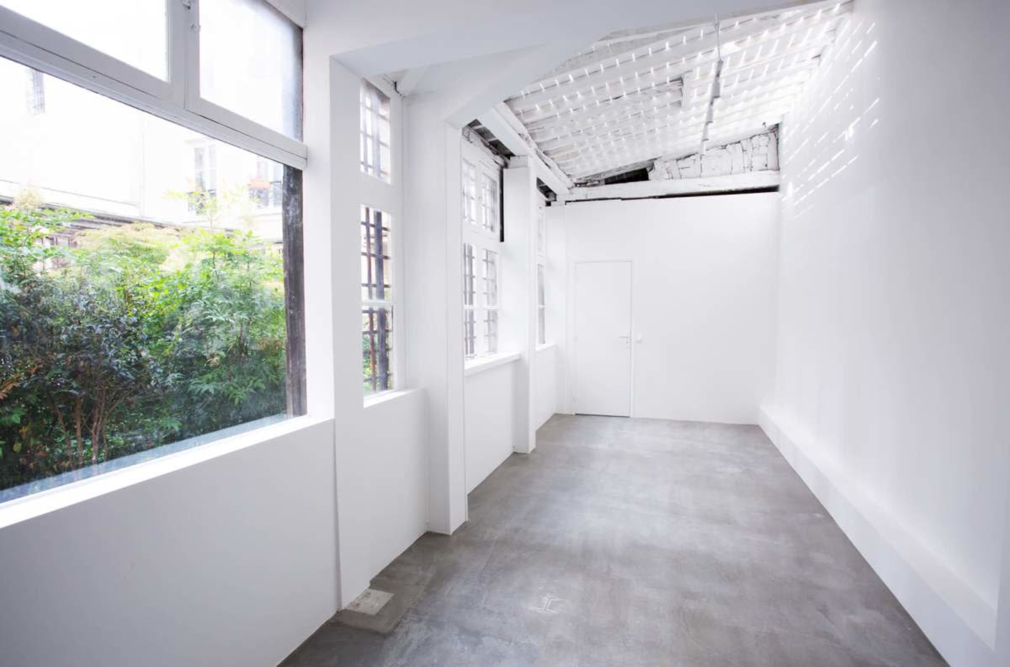 The image shows a spacious, bright room with white walls, large windows, and a concrete floor.