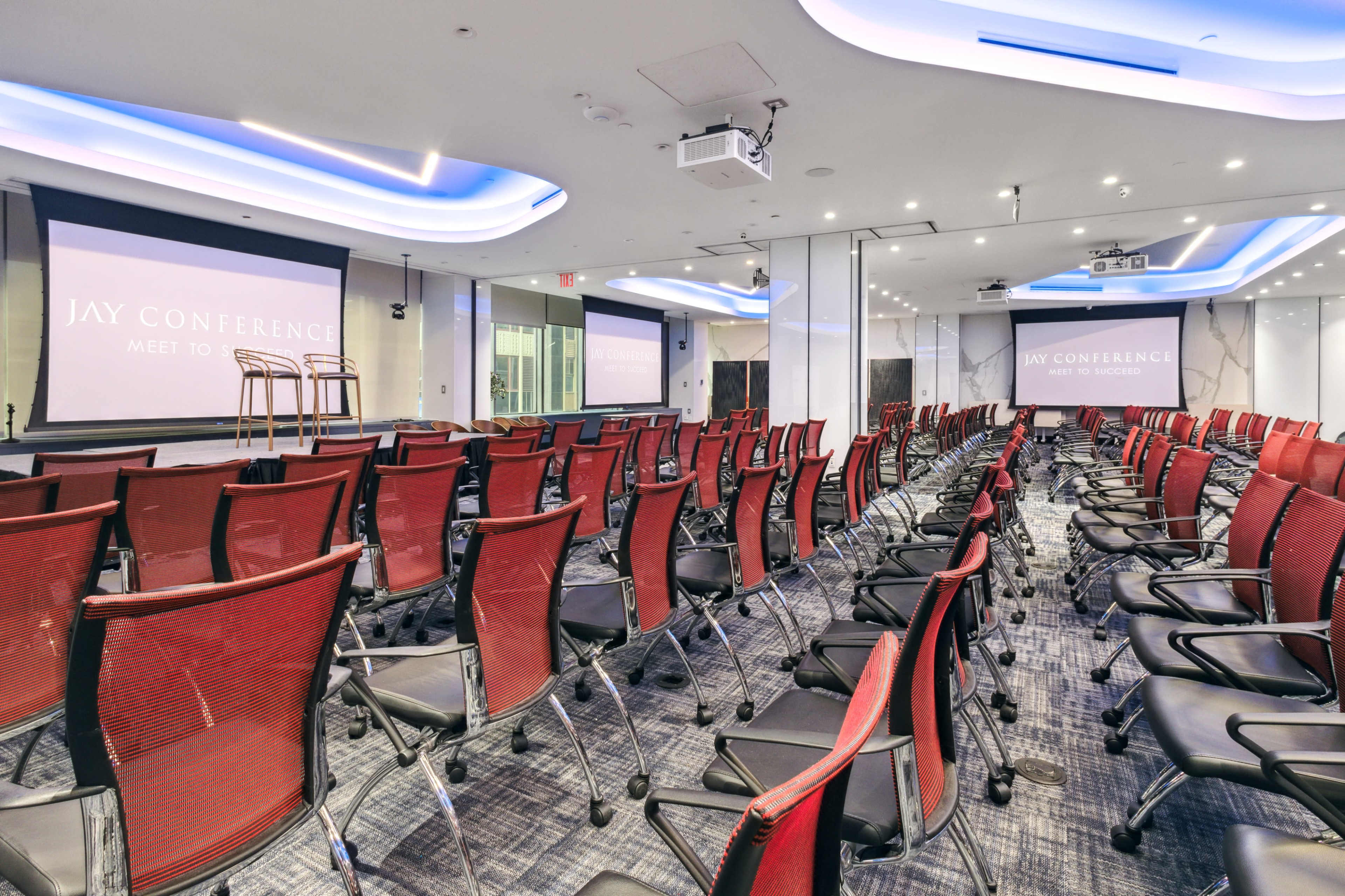 A conference room set up with rows of red chairs facing a stage, with screens displaying "JAY CONFERENCE" in the background.
