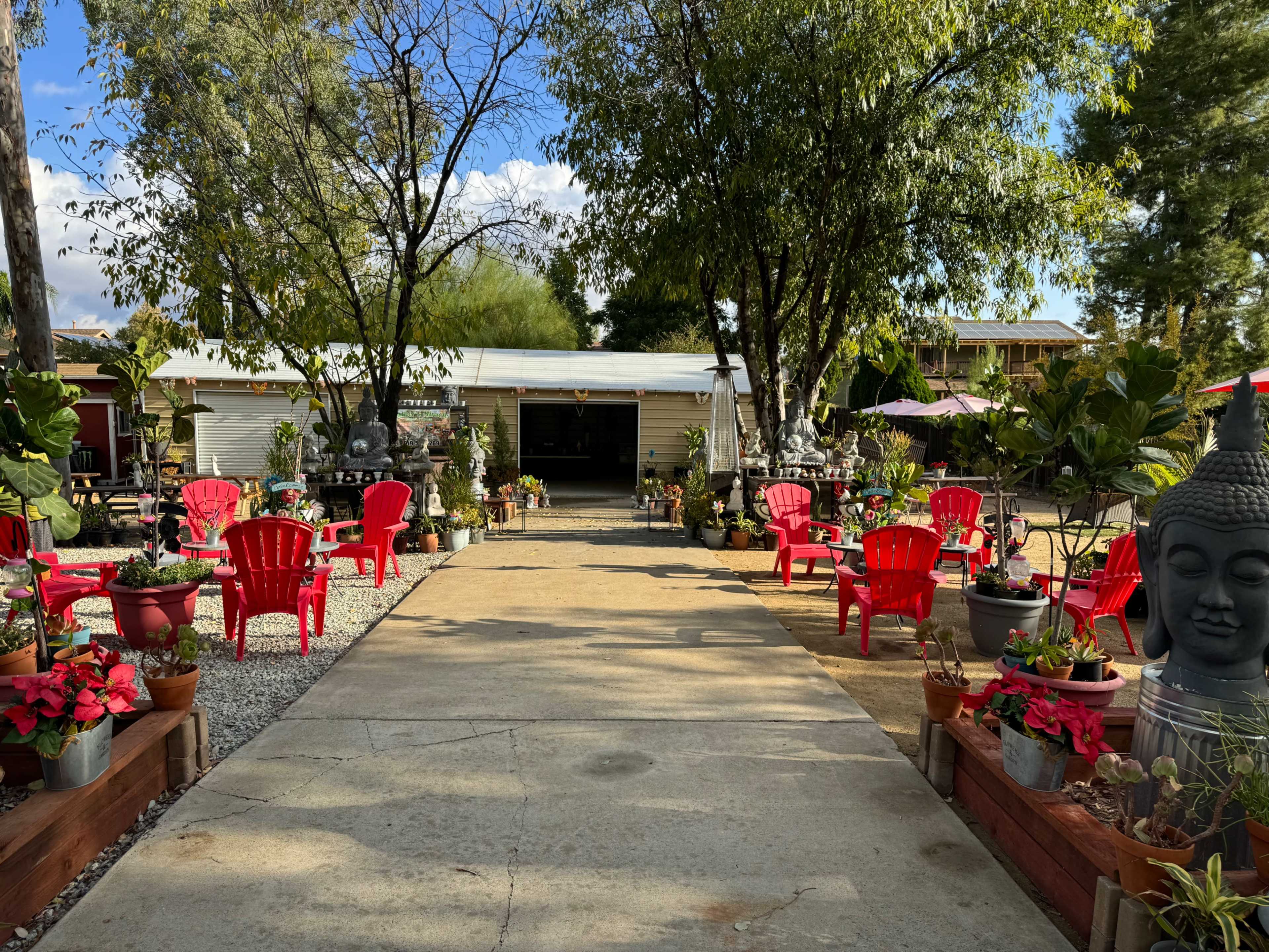 A pathway leads through a garden filled with red chairs, potted plants, and stone sculptures, framed by trees on either side.