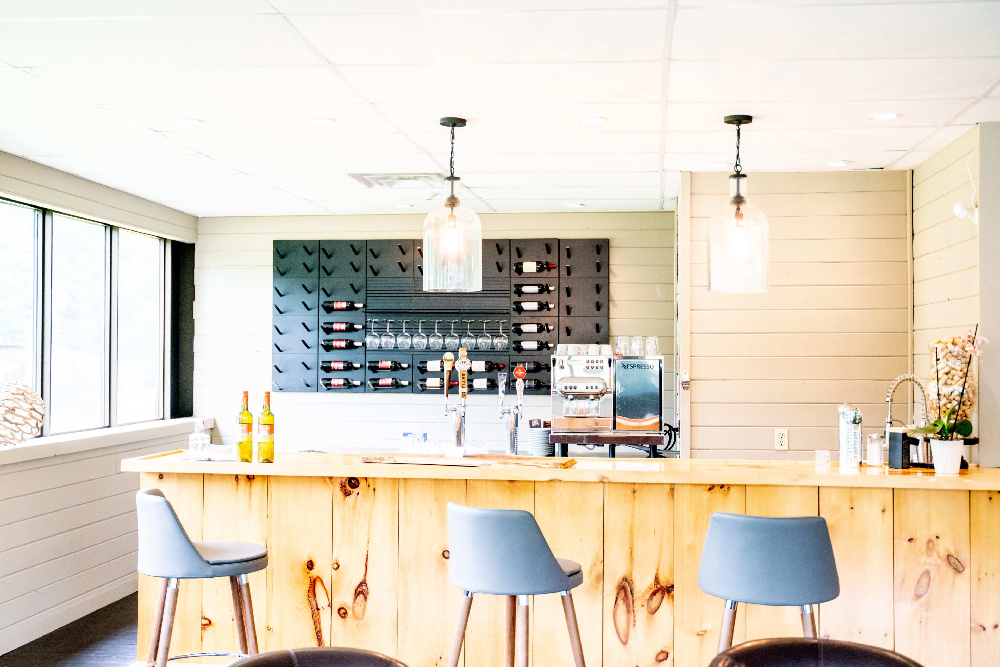 A modern bar area with a wooden counter, three gray bar stools, and a wall-mounted wine rack holding several bottles.