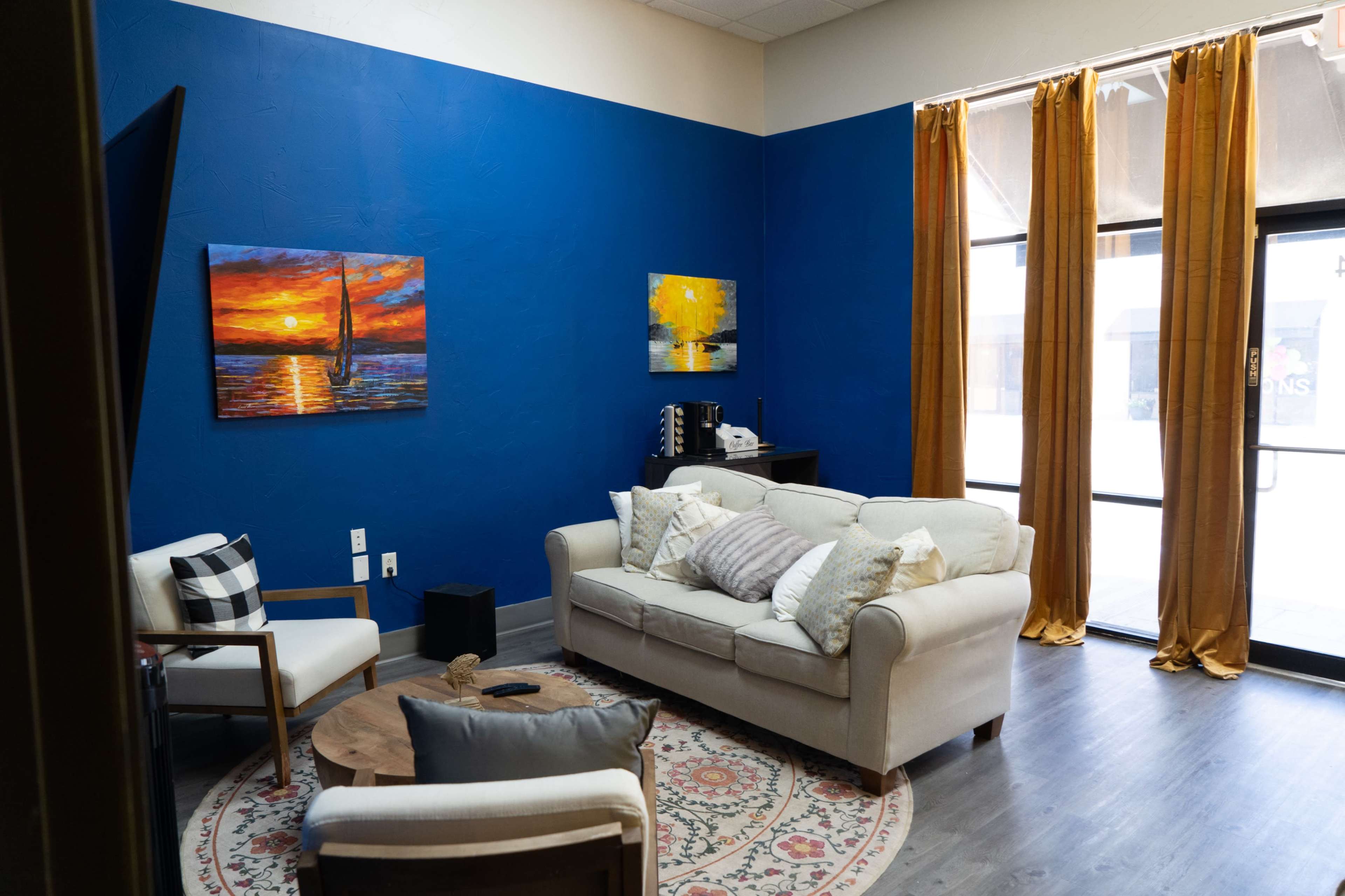 A cozy lounge area with a light-colored sofa, a round wooden coffee table, and two paintings on the blue wall, illuminated by natural light coming through large windows.