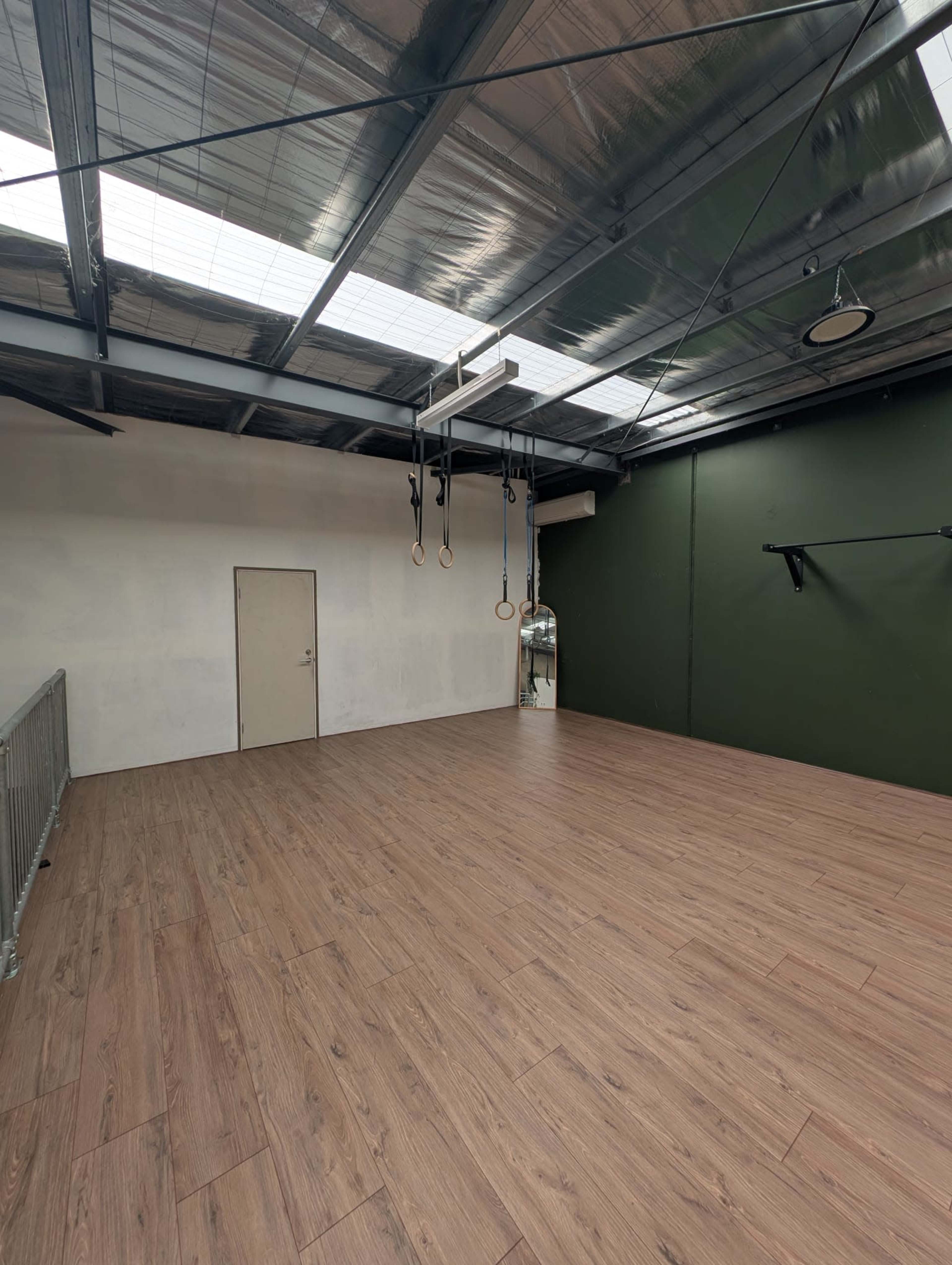 The image shows an empty gym space with wooden flooring, a green wall, and gymnastic rings hanging from the ceiling.