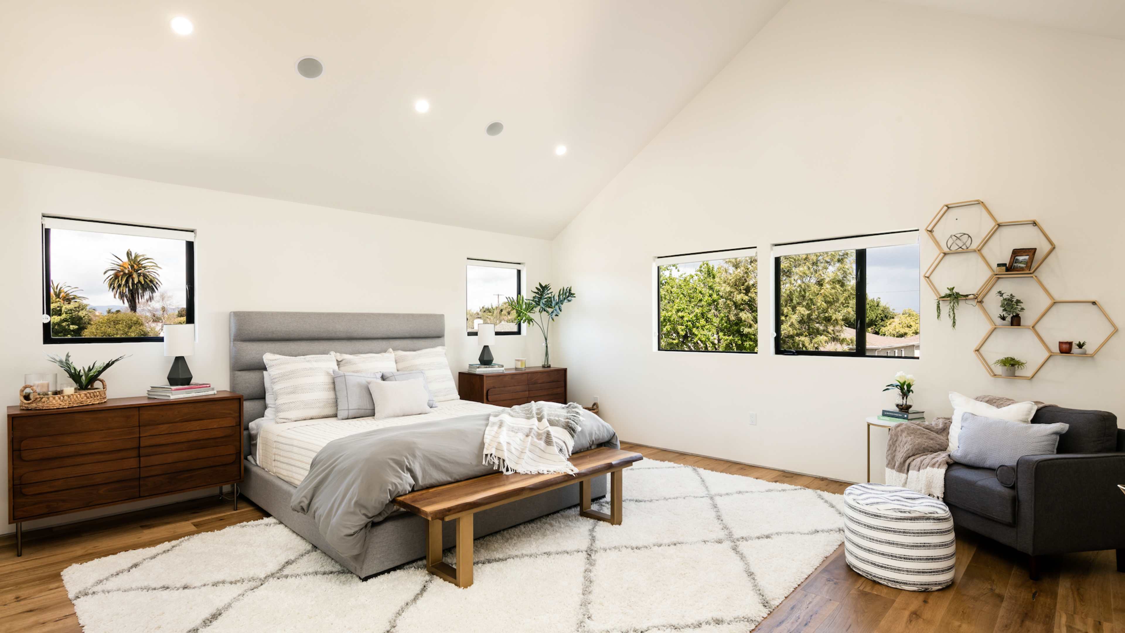 The image shows a modern bedroom with a large bed, wooden furniture, and multiple windows allowing natural light to enter the space.