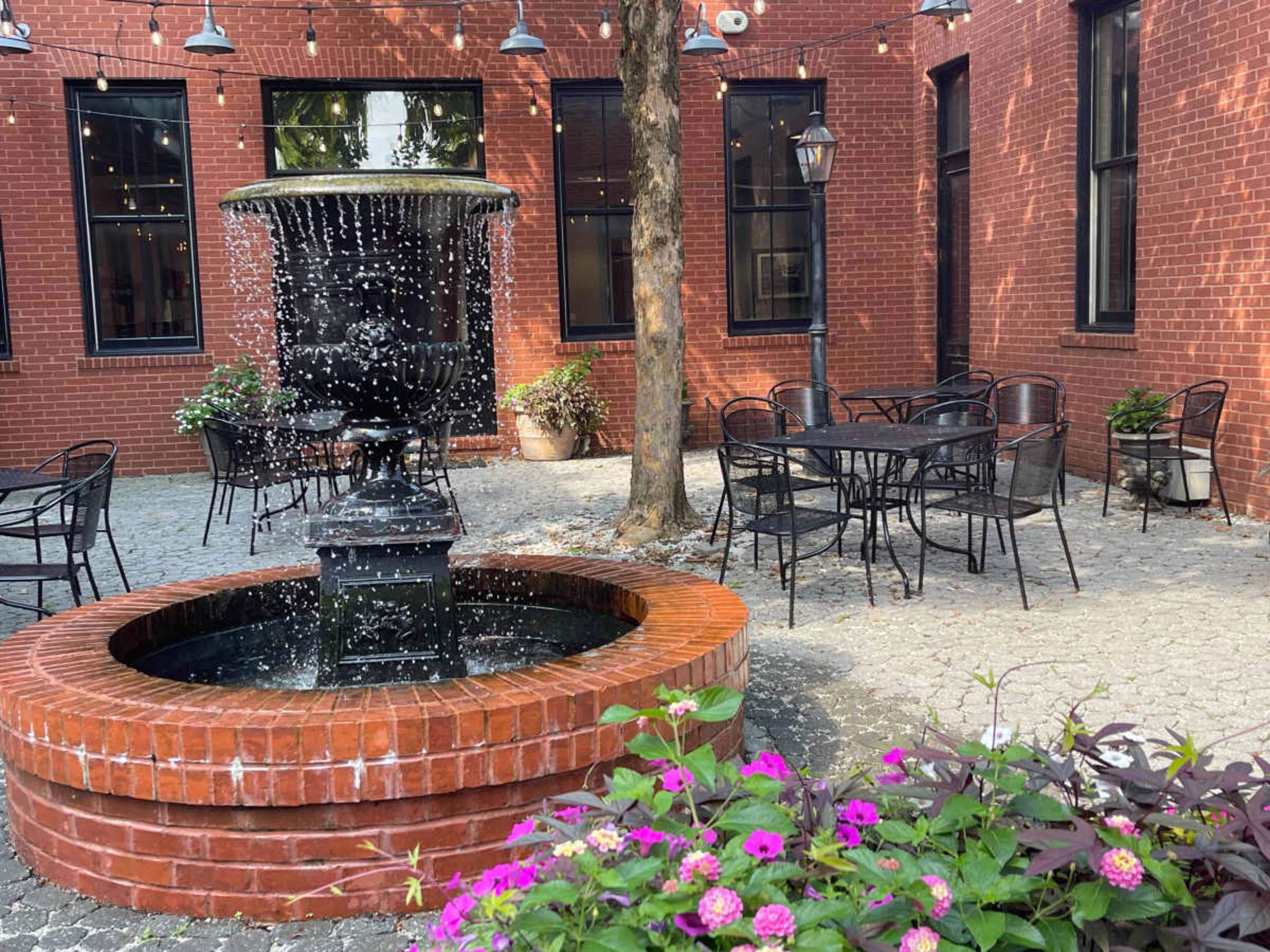 The image shows a courtyard featuring a brick fountain, surrounded by tables and chairs, with potted plants and string lights overhead.