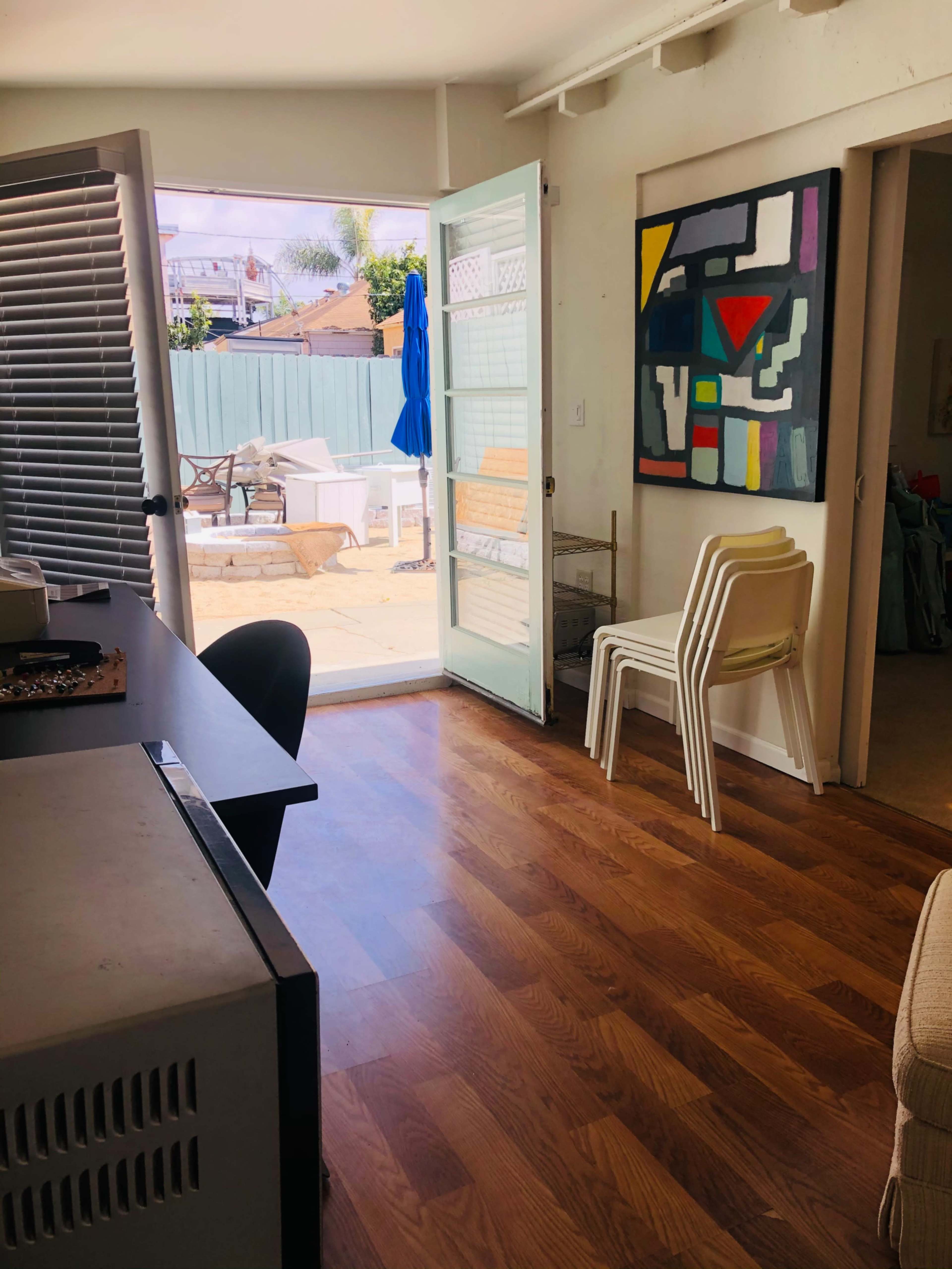 A room with hardwood floors features a desk and several stacked chairs, leading to a patio area visible through an open sliding glass door.