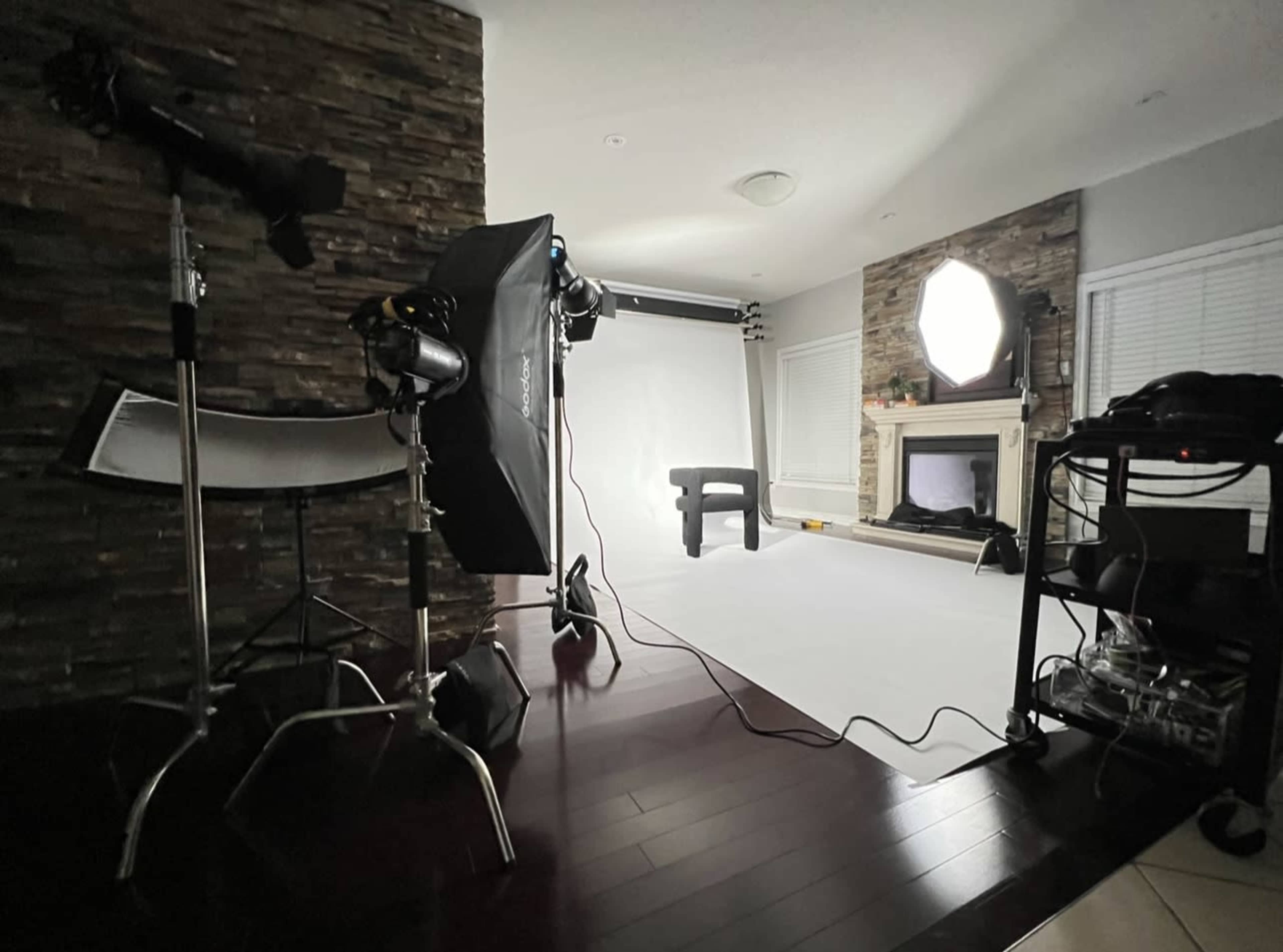 The image shows a photography studio setup with lighting equipment, a backdrop, and a wooden stool in a room with stone accents and hardwood flooring.