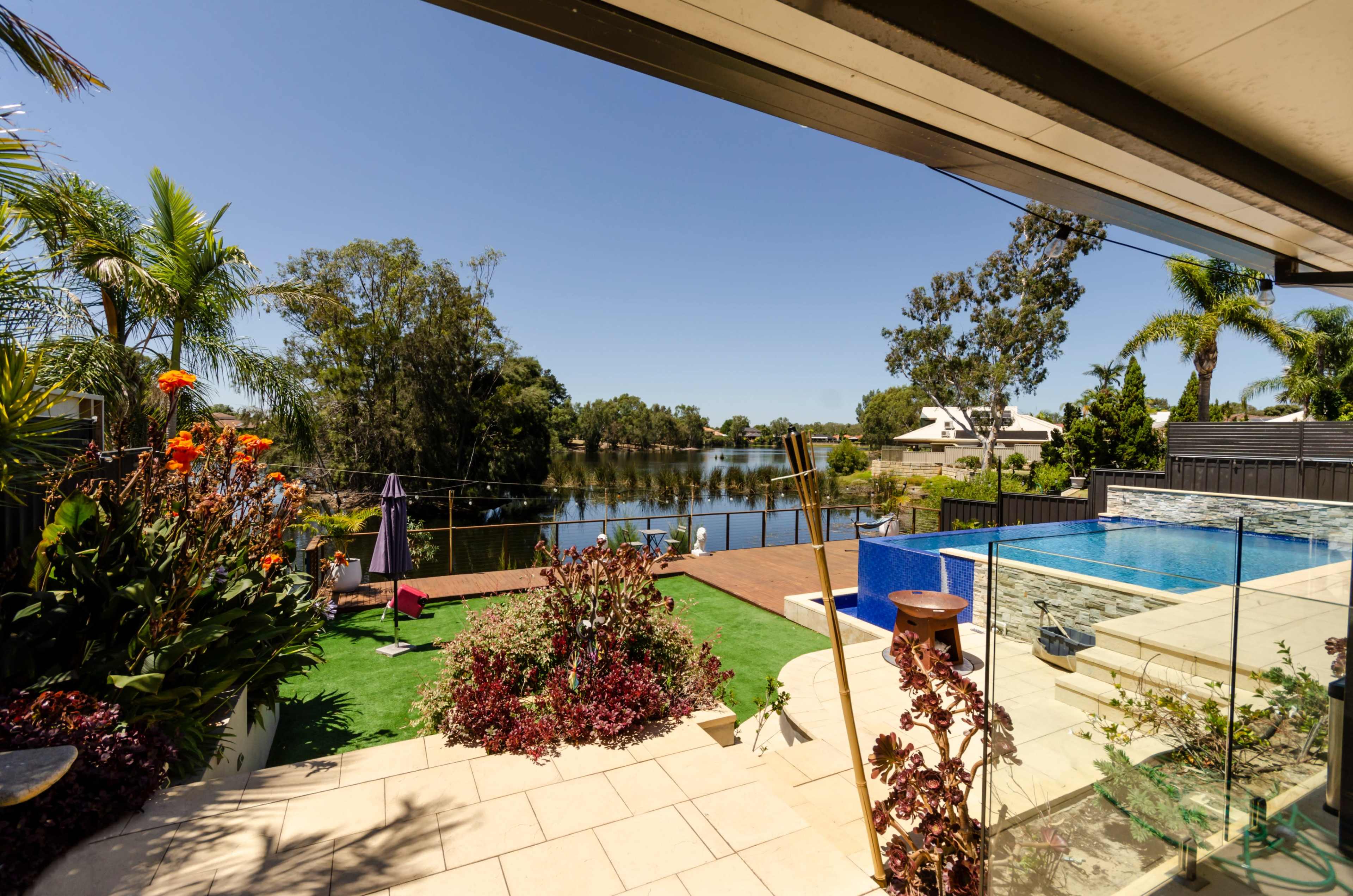 The image shows a backyard with a swimming pool, landscaped gardens, and a view of a calm water body surrounded by trees.