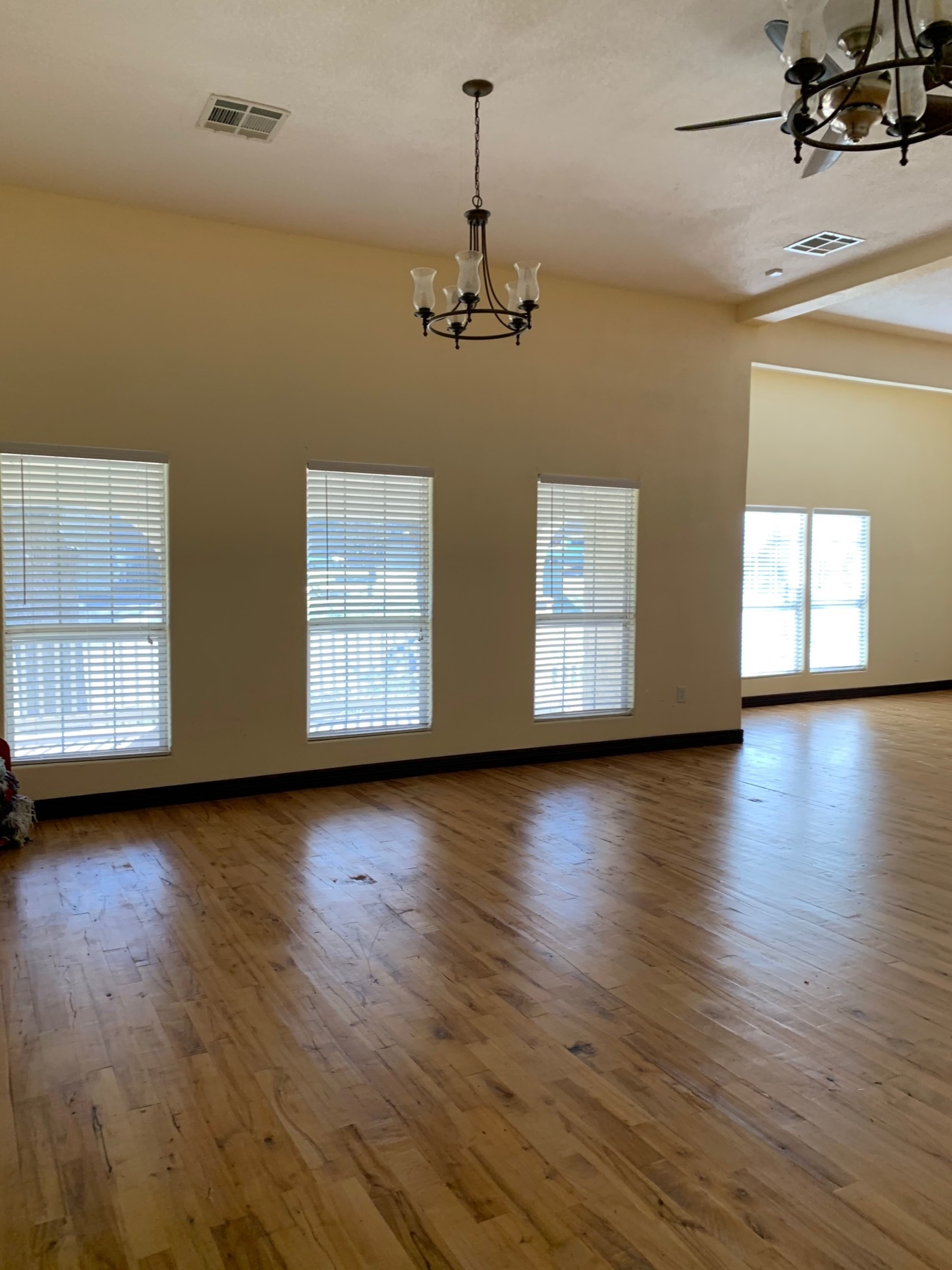 A spacious room with wooden floors features several windows adorned with blinds and a chandelier overhead.